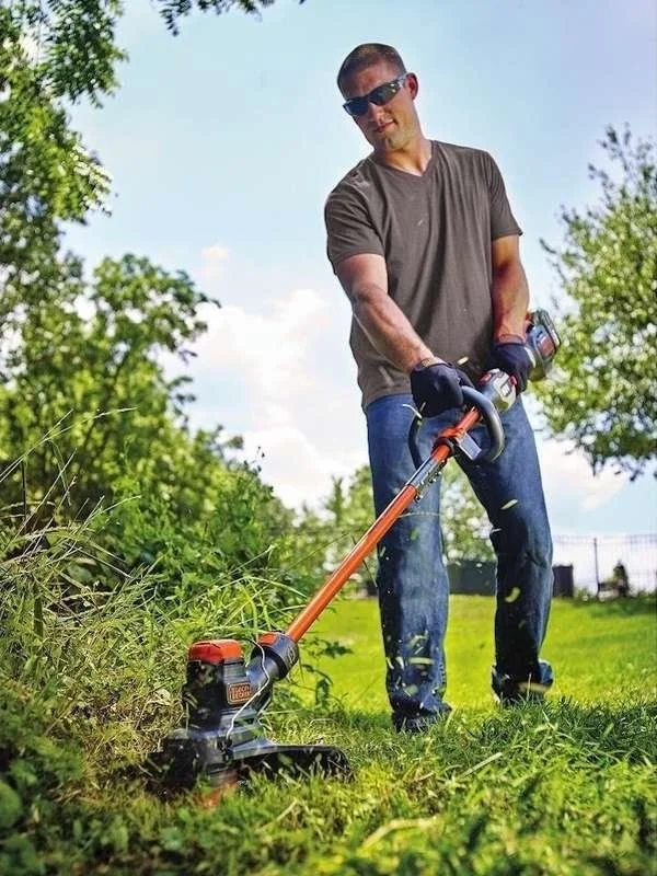 Man wearing sunglasses and a gray t-shirt using a weed trimmer to cut grass outdoors on a sunny day.