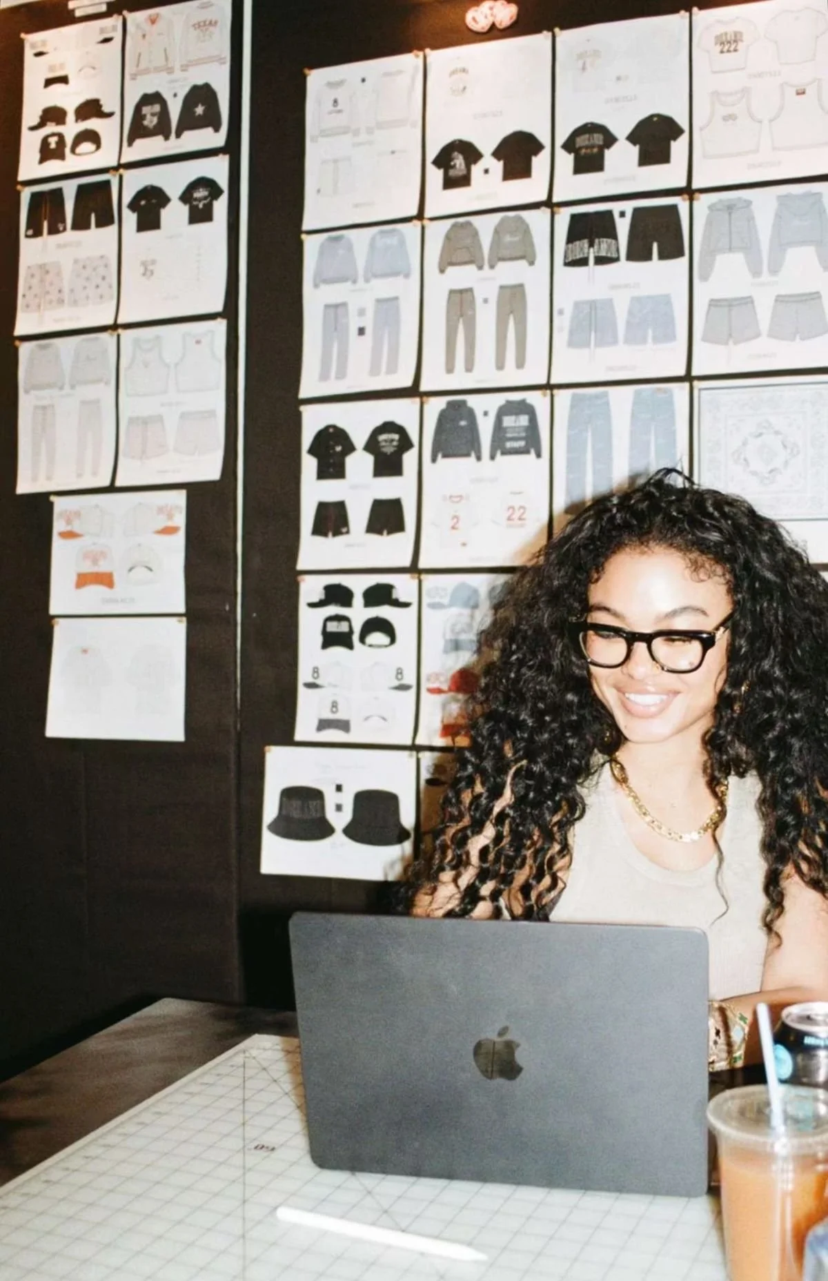 A woman with curly hair and glasses sitting at a table with a MacBook, in front of a wall covered with clothing sketches and design sketches.