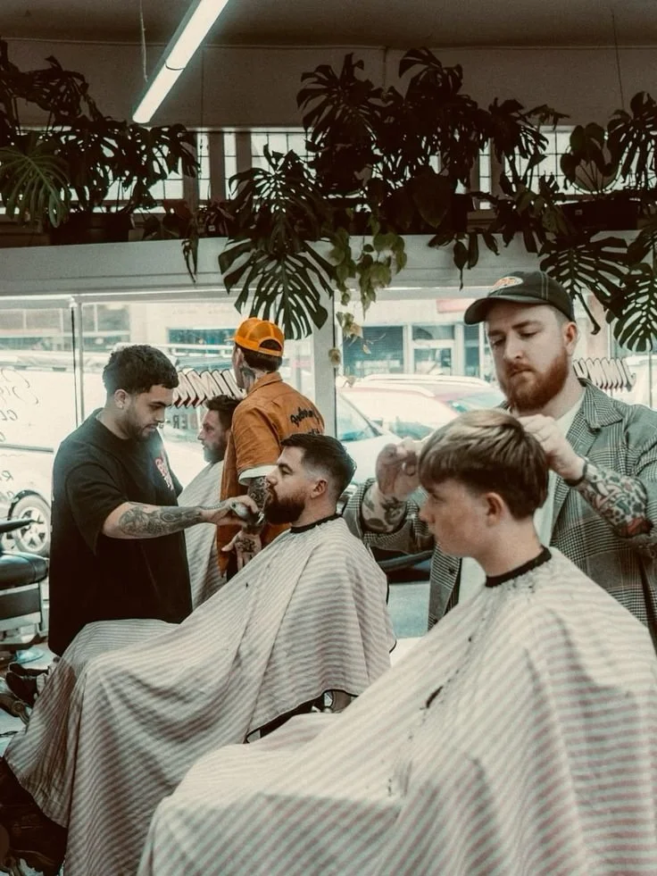 People getting haircuts at a barbershop with large indoor plants and a city view outside the window.