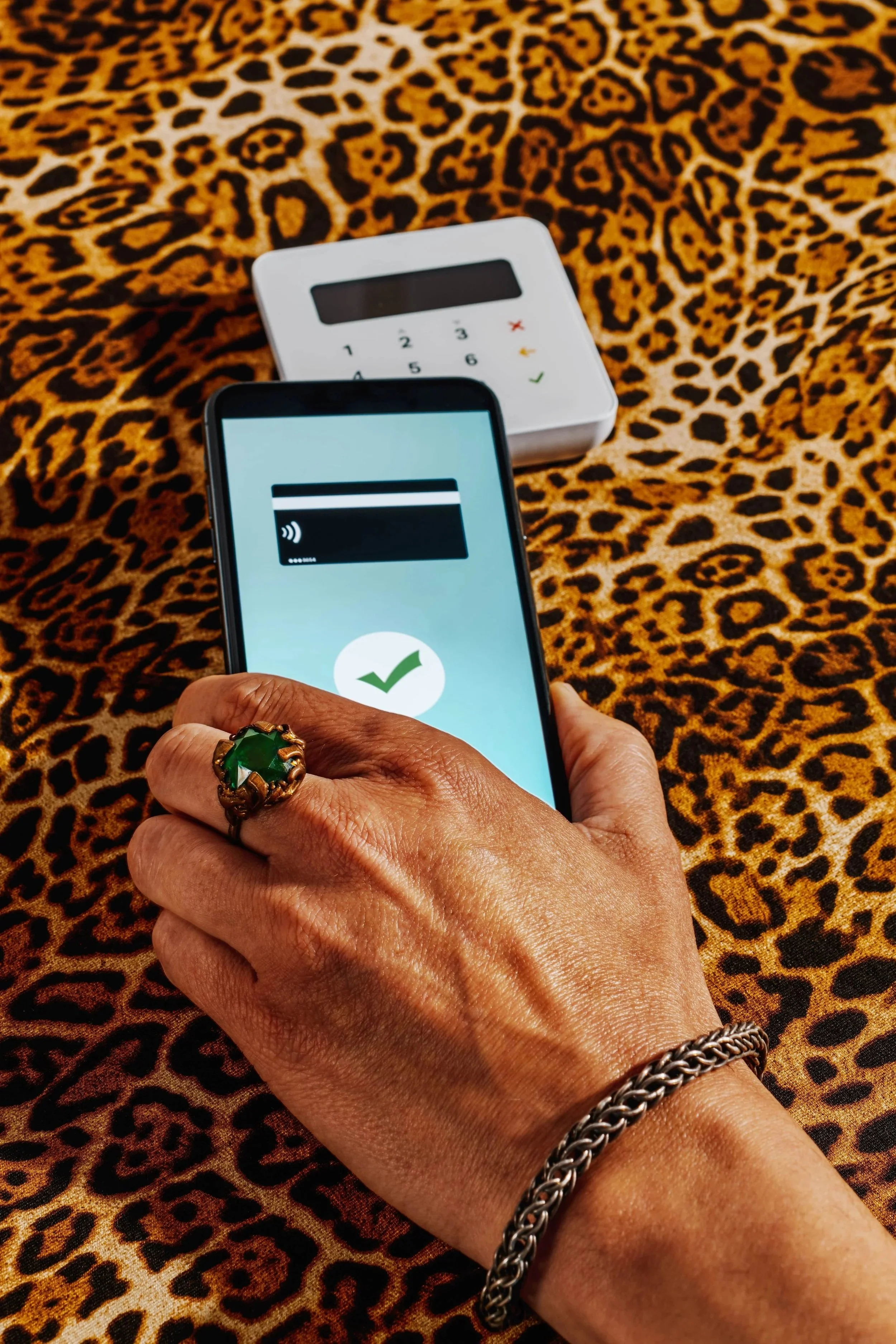 Close-up of a hand holding a smartphone with a contactless payment screen, in front of a leopard print fabric background. In the background, there is a small white device, possibly a card reader.