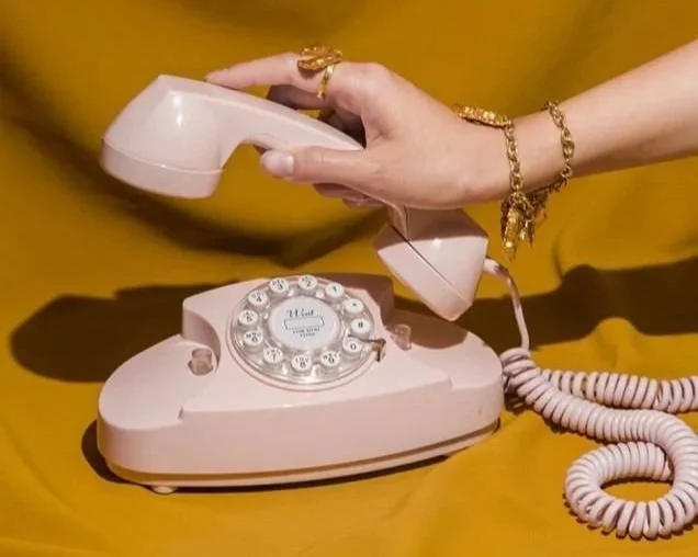 A person holding a pink rotary dial phone with a coiled cord against a yellow background.