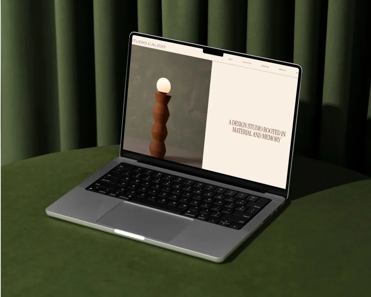 A laptop on a green table with a dark green curtain in the background. The screen shows a minimalist webpage with a photo of a wooden sculpture topped with a white sphere and text that reads, "A design studio rooted in material and memory."