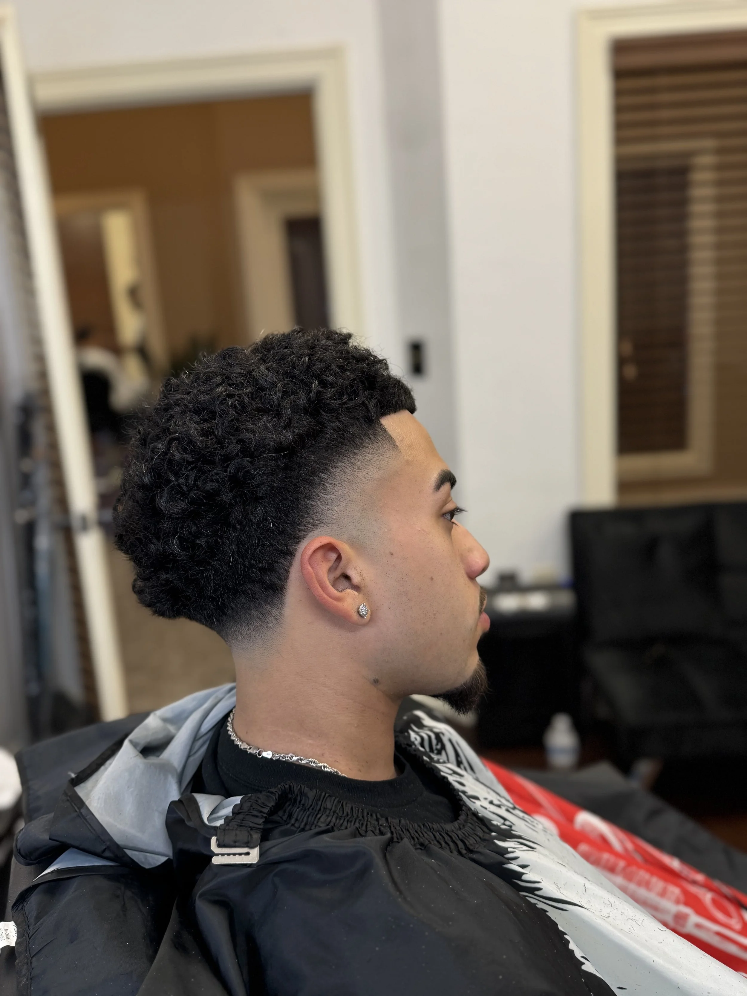 Side view of a young man with a curly high fade hairstyle, wearing diamond earrings, a chain necklace, and a black shirt, sitting in a barber shop chair.