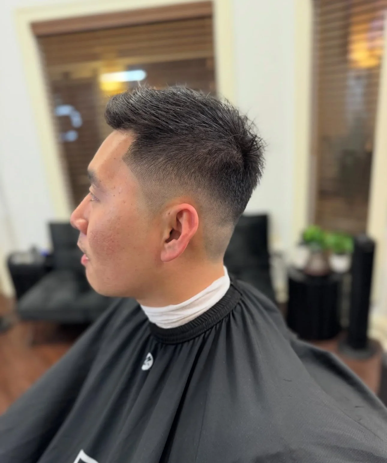 A man with a fresh men's haircut featuring a high fade and textured top, sitting in a barber's chair in a salon with wooden window blinds and plants in the background.