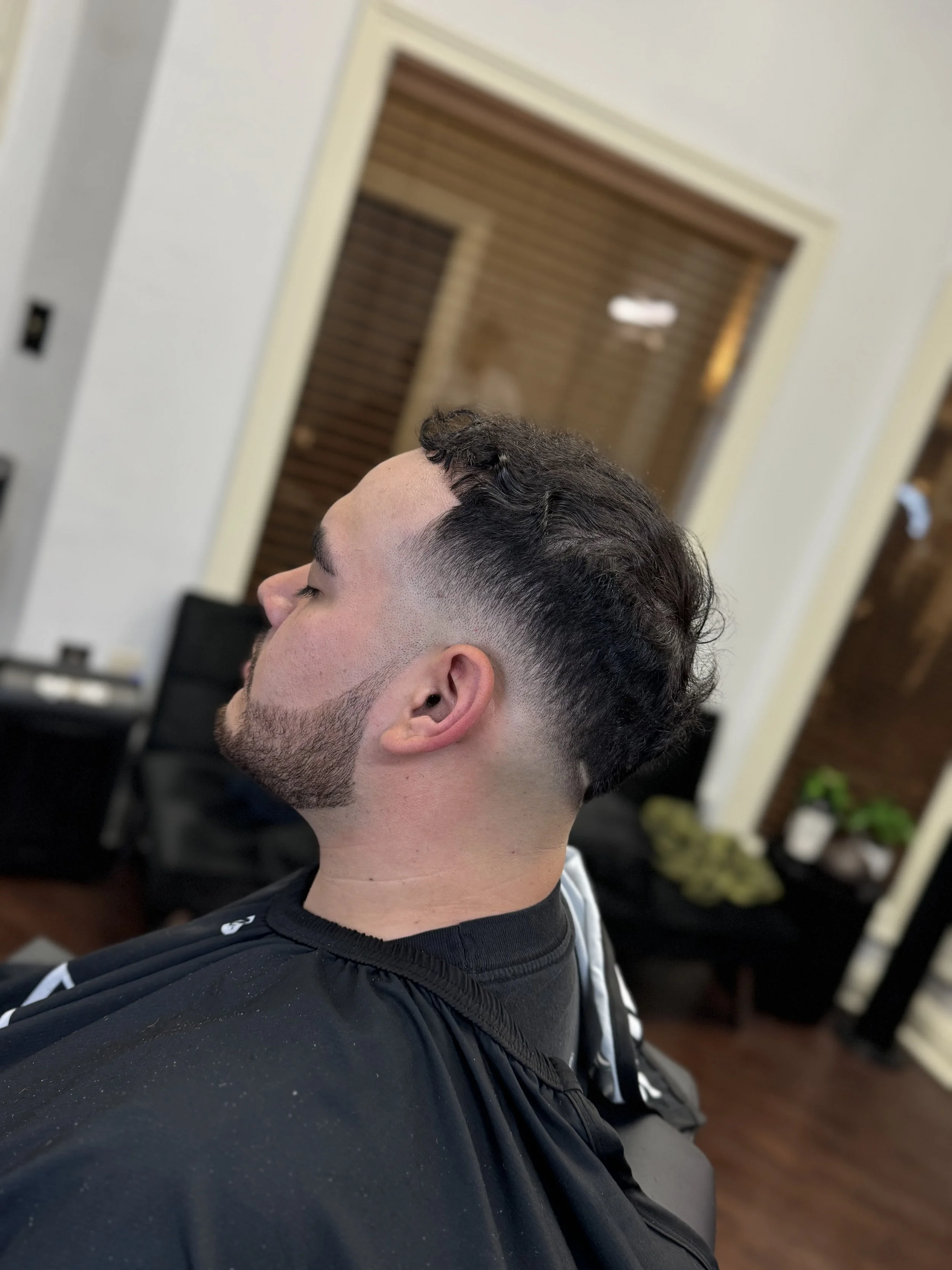 Young man with a fresh fade haircut relaxing in a barbershop chair, eyes closed, wearing a black barber cape, inside a modern barbershop with wood and white decor, plants, and chairs in the background.
