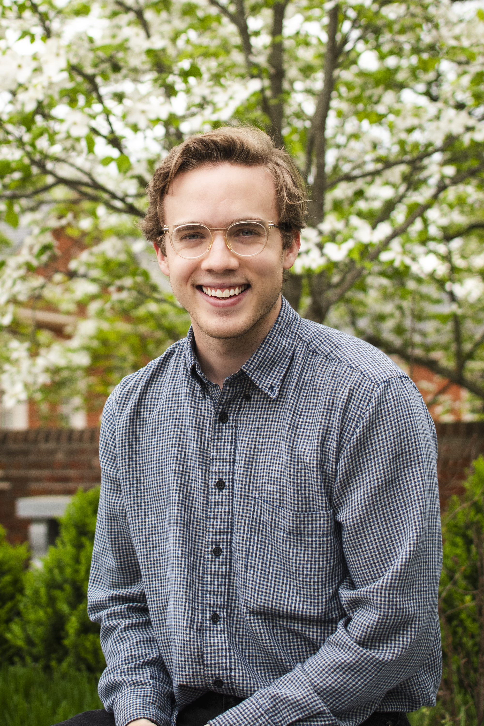 A young man with glasses, light brown hair, and a checkered shirt smiling while sitting outdoors in front of a blooming tree with white flowers and green leaves.