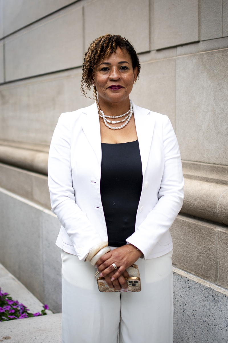 A woman standing outdoors in front of a beige stone wall, wearing a white blazer over a black top, accessorized with multiple pearl necklaces, bracelets, and a ring, holding a patterned phone case.