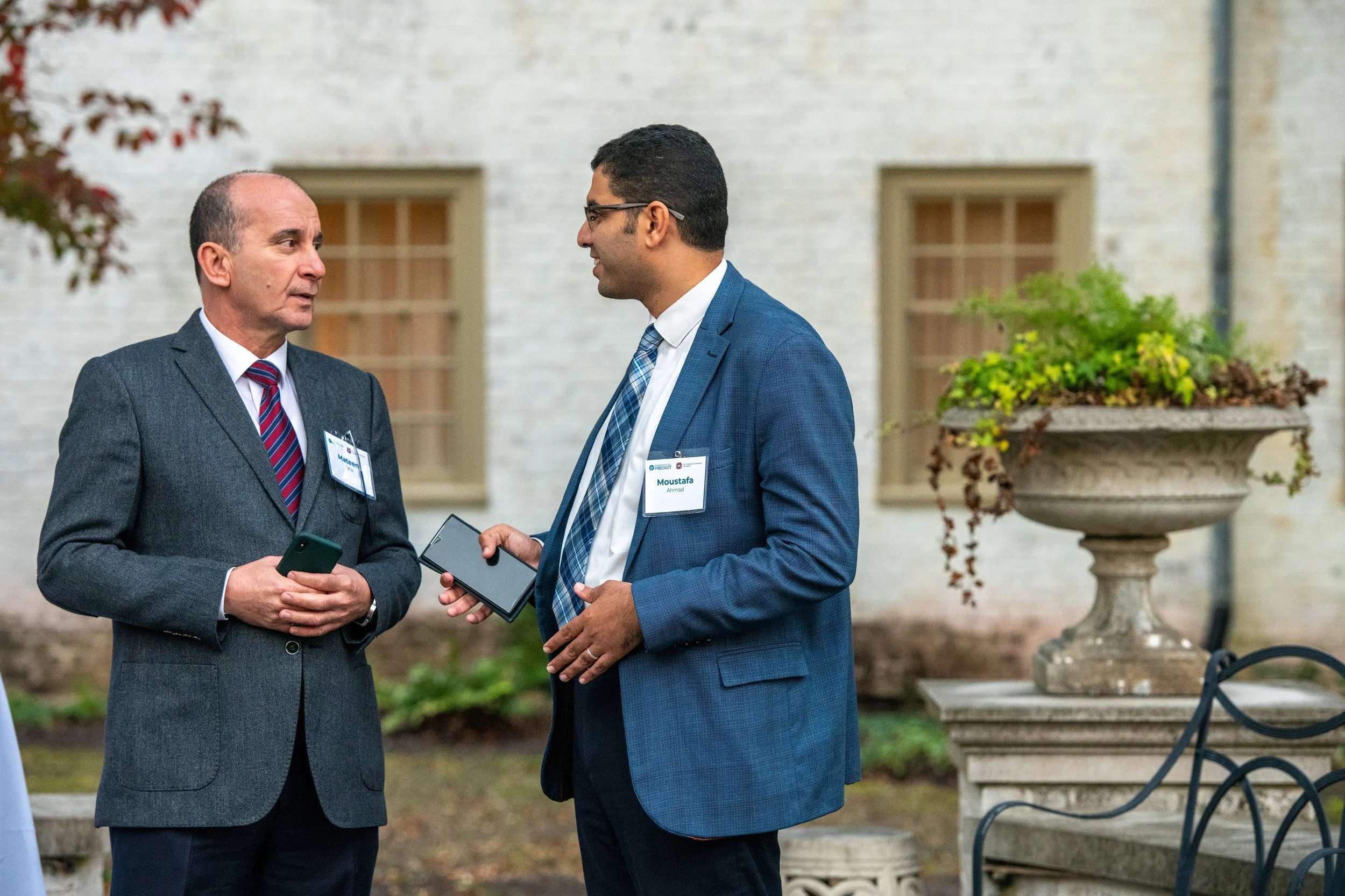 Two men in suits having a conversation outdoors next to a decorative stone planter with plants, with a background of a building with two windows.