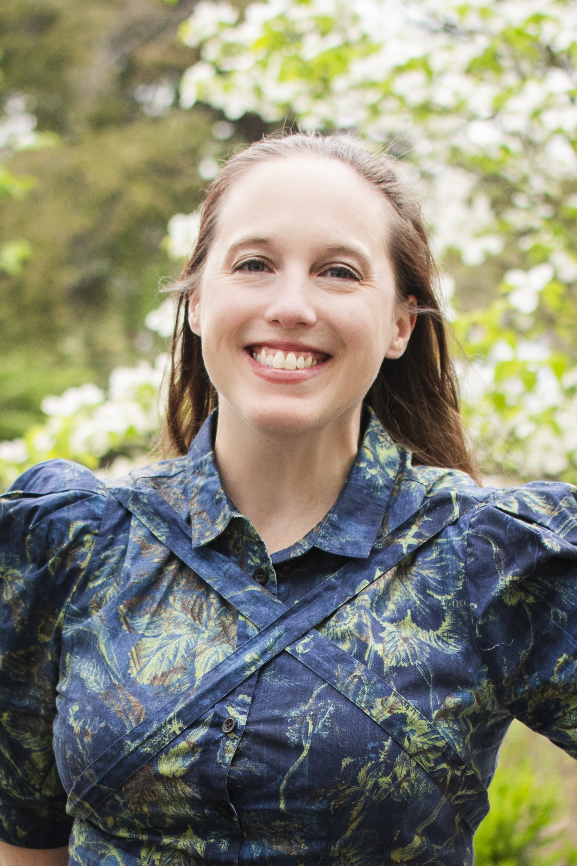 A woman smiling outdoors in front of flowering trees, wearing a blue floral patterned shirt.