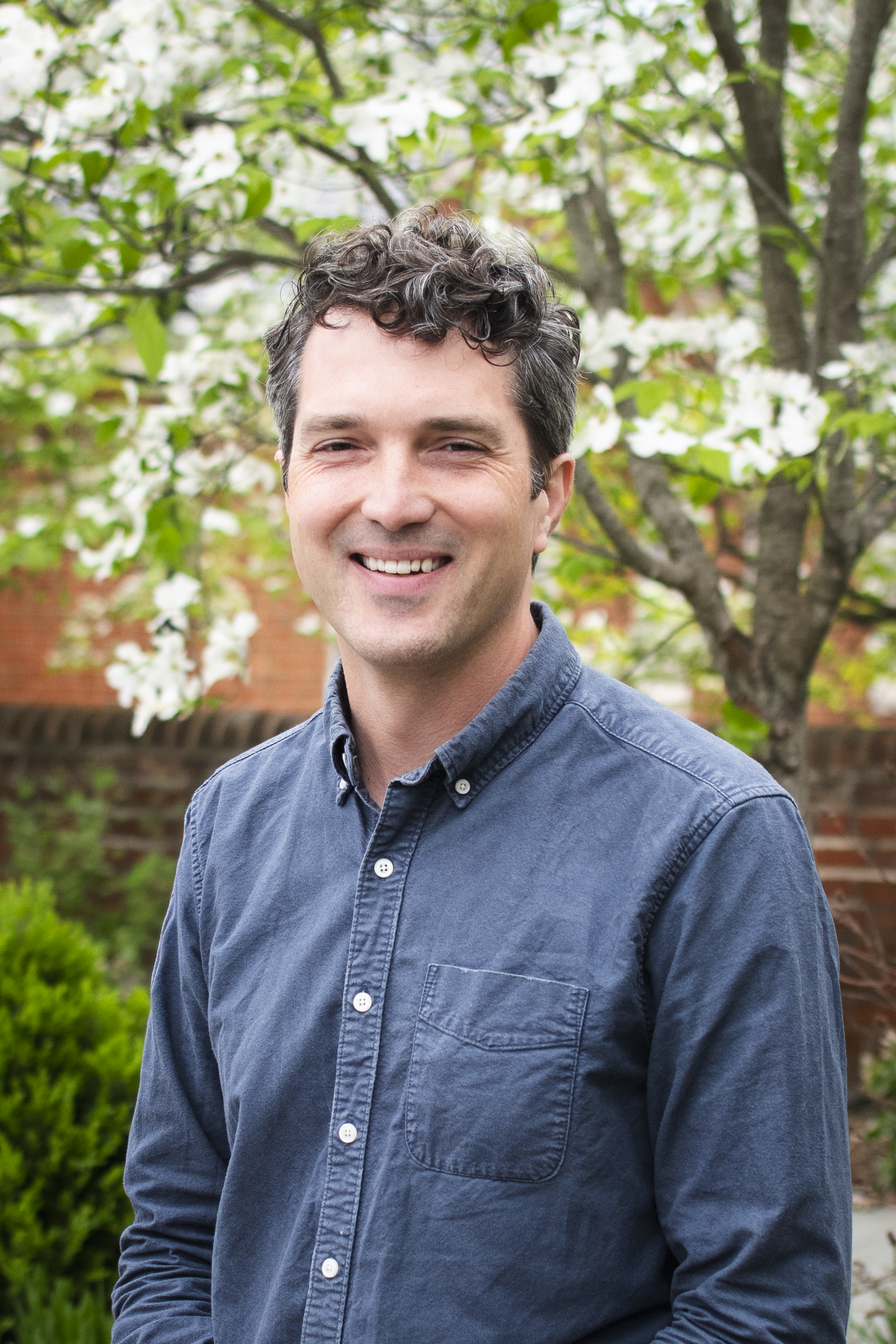 A man with dark hair, wearing a blue button-up shirt, smiling outdoors, with a background of green trees and white blossoms.
