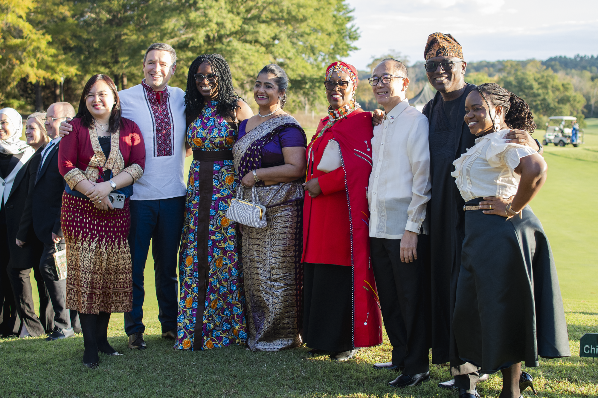 A diverse group of men and women standing outdoors on a sunny day, wearing traditional and modern clothing, smiling and posing for a photo.