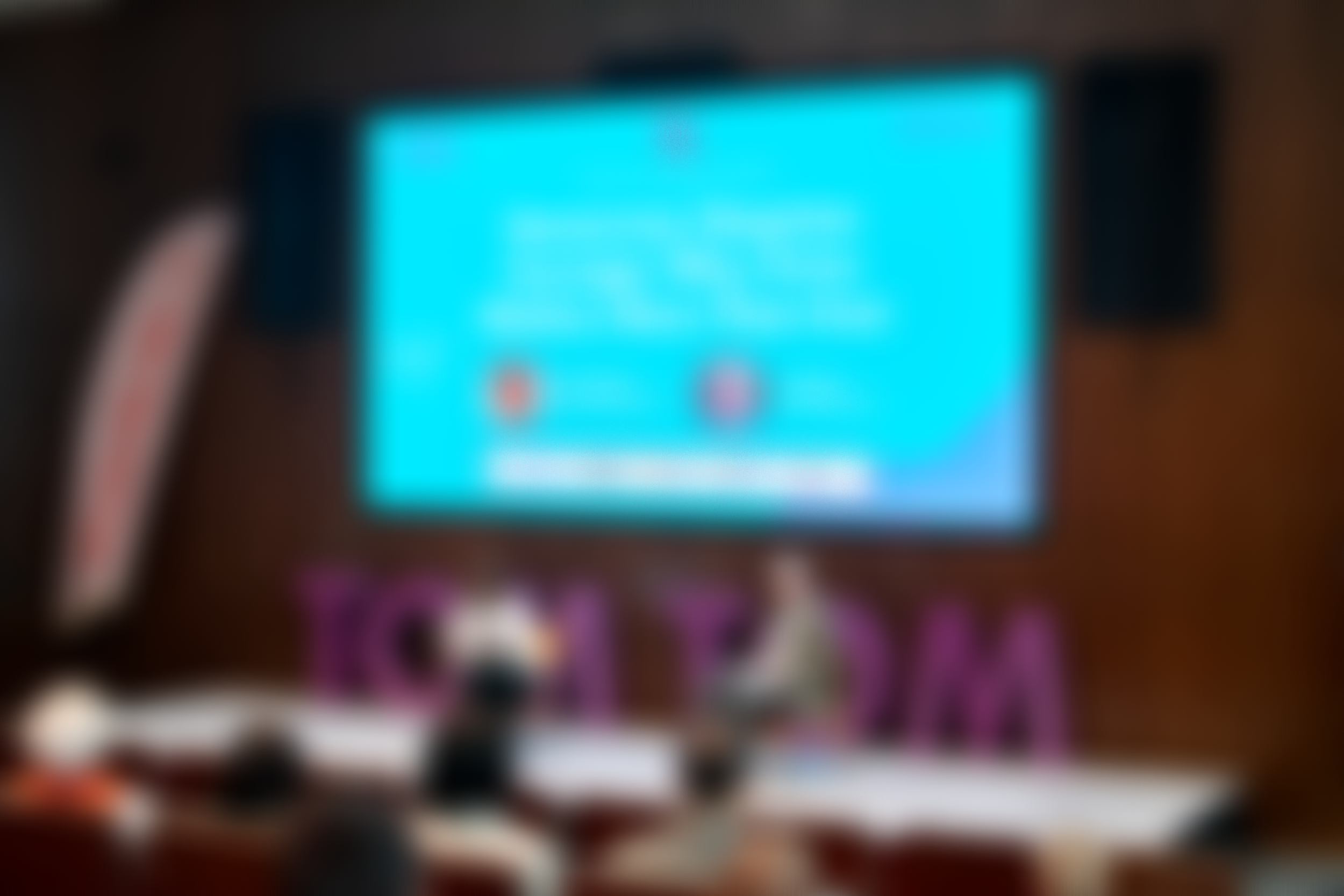A blurry image of two people conversing on stage, under a bright screen.