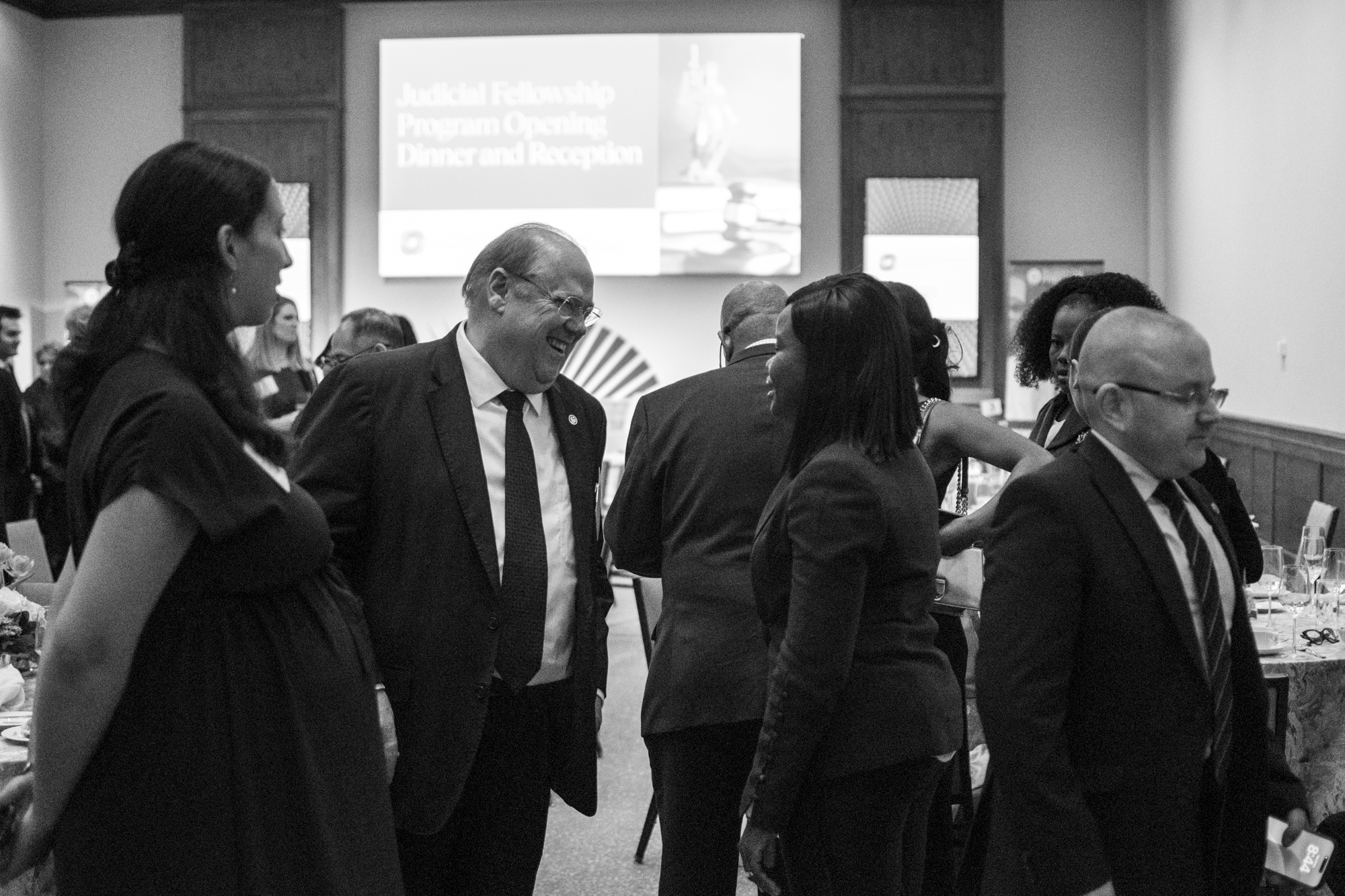 A group of people at a formal event, dressed in suits and dresses, engaged in conversation, with a presentation slide in the background reading 'Judicial Fellowship Program Opening Dinner and Reception'.
