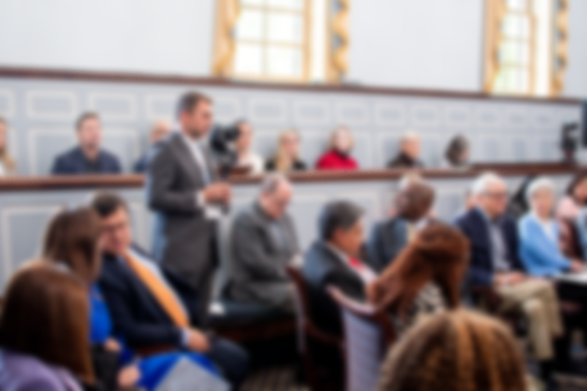 A man in a suit speaking or presenting in a conference room filled with seated attendees.