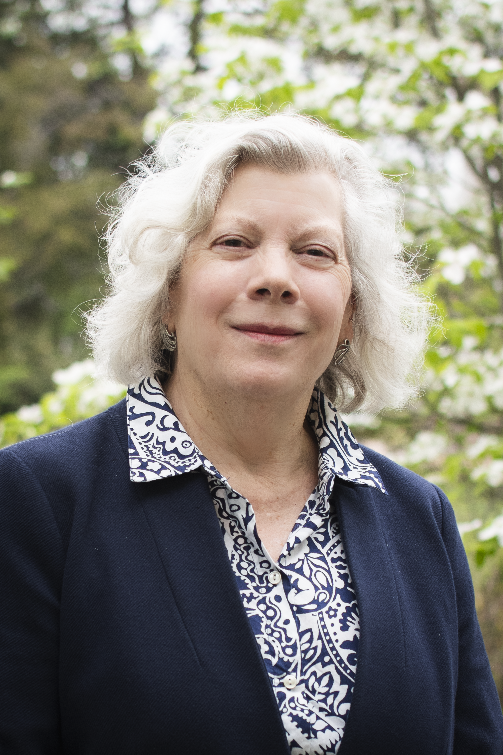 Close-up of a smiling elderly woman with white hair outdoors, wearing a navy blazer and a patterned blouse against a background of green foliage.