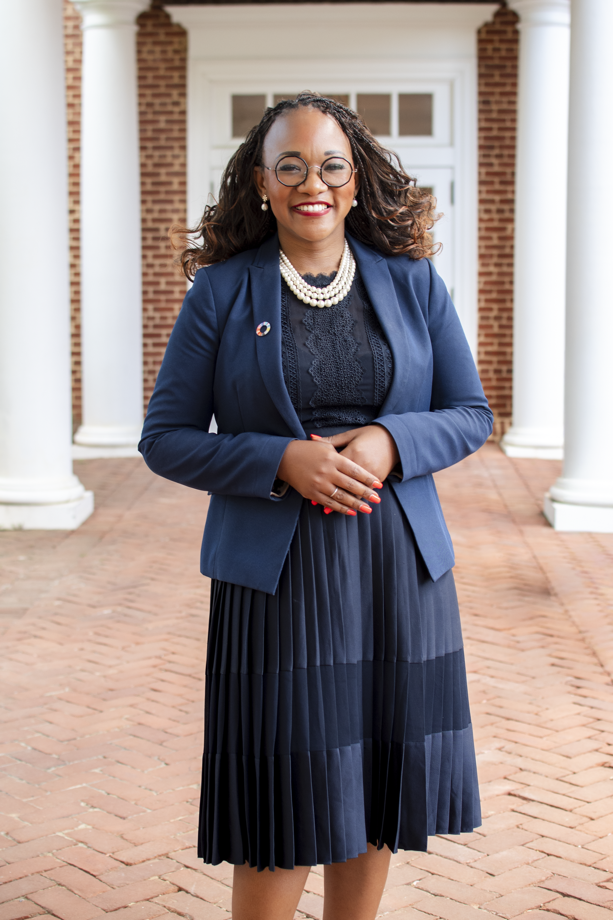 A woman with glasses and pearl earrings smiling outdoors, dressed in a navy blue blazer and pleated skirt, standing in front of a white columned building with a brick pathway.