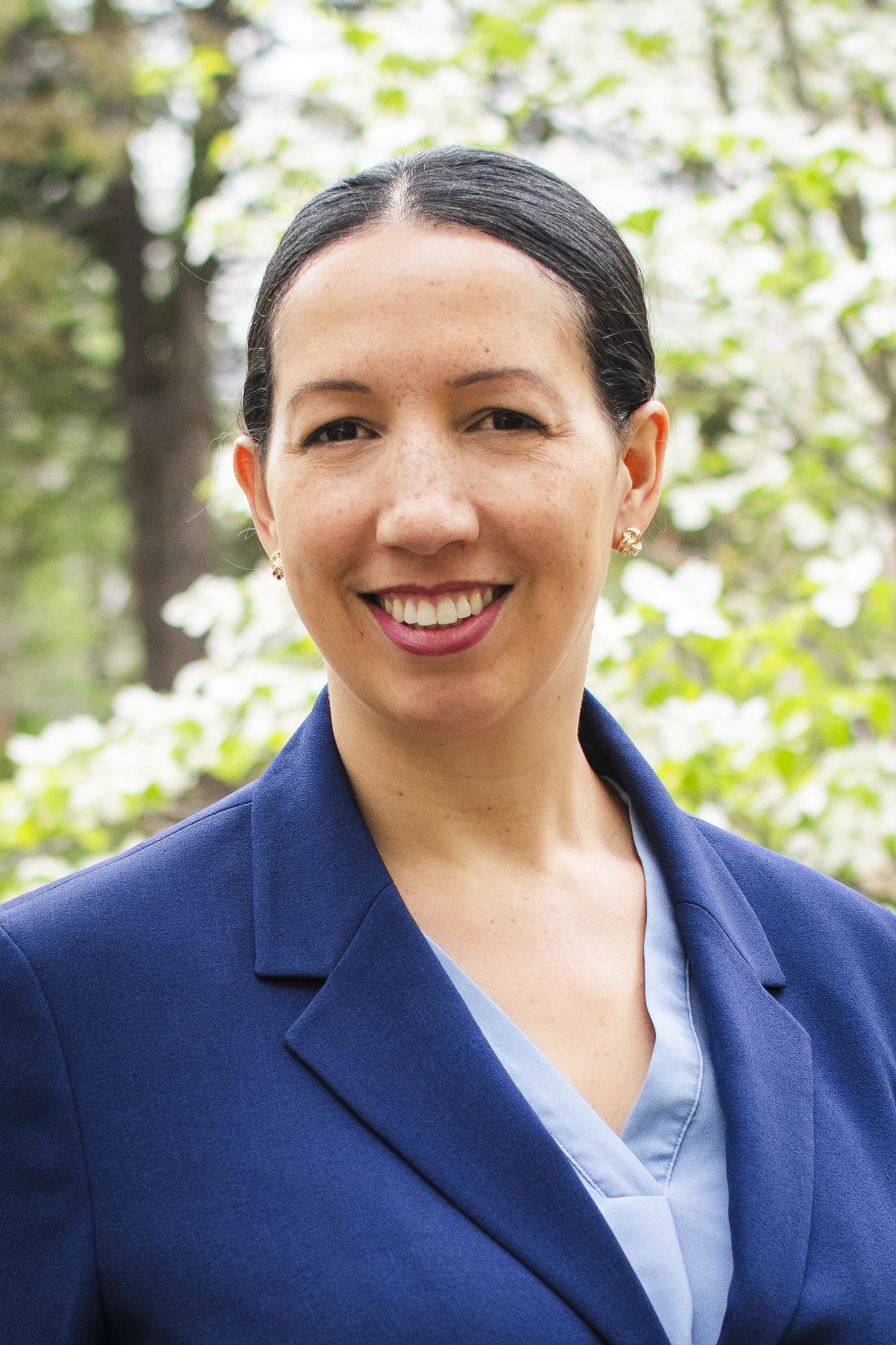 Close-up of a woman smiling outdoors, wearing a navy blazer and blue top, with green foliage in the background.