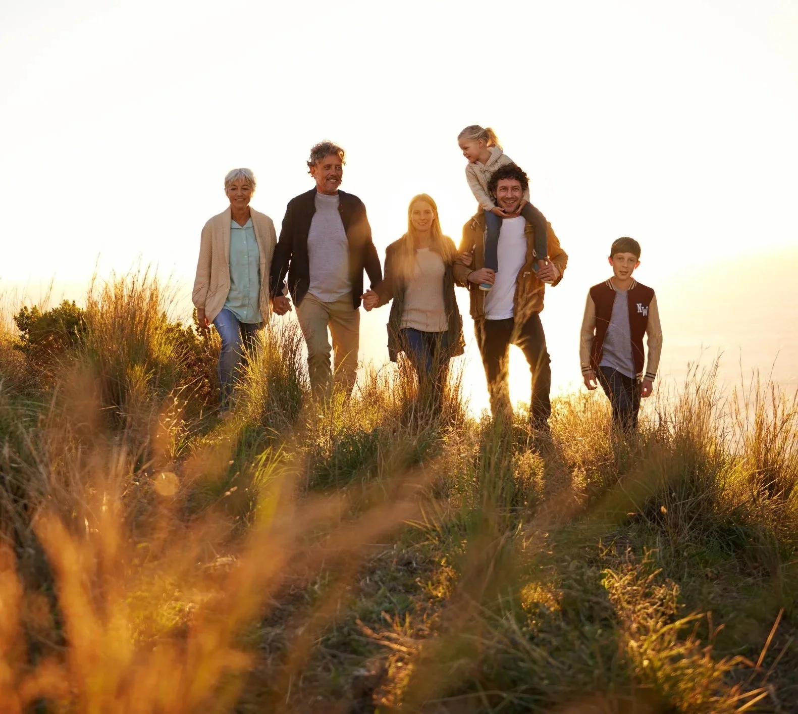 A happy multi-generational family walking together in a grassy field during sunset, with two children sitting on adults' shoulders.