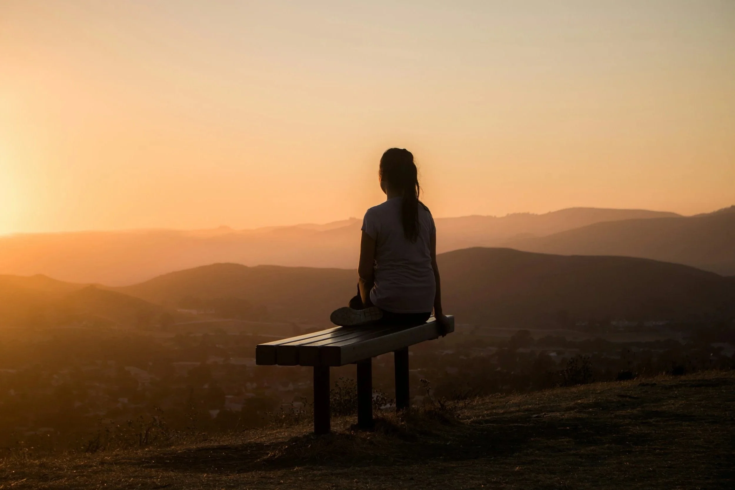 A girl sitting on a park bench overlooking a sunset over rolling hills.