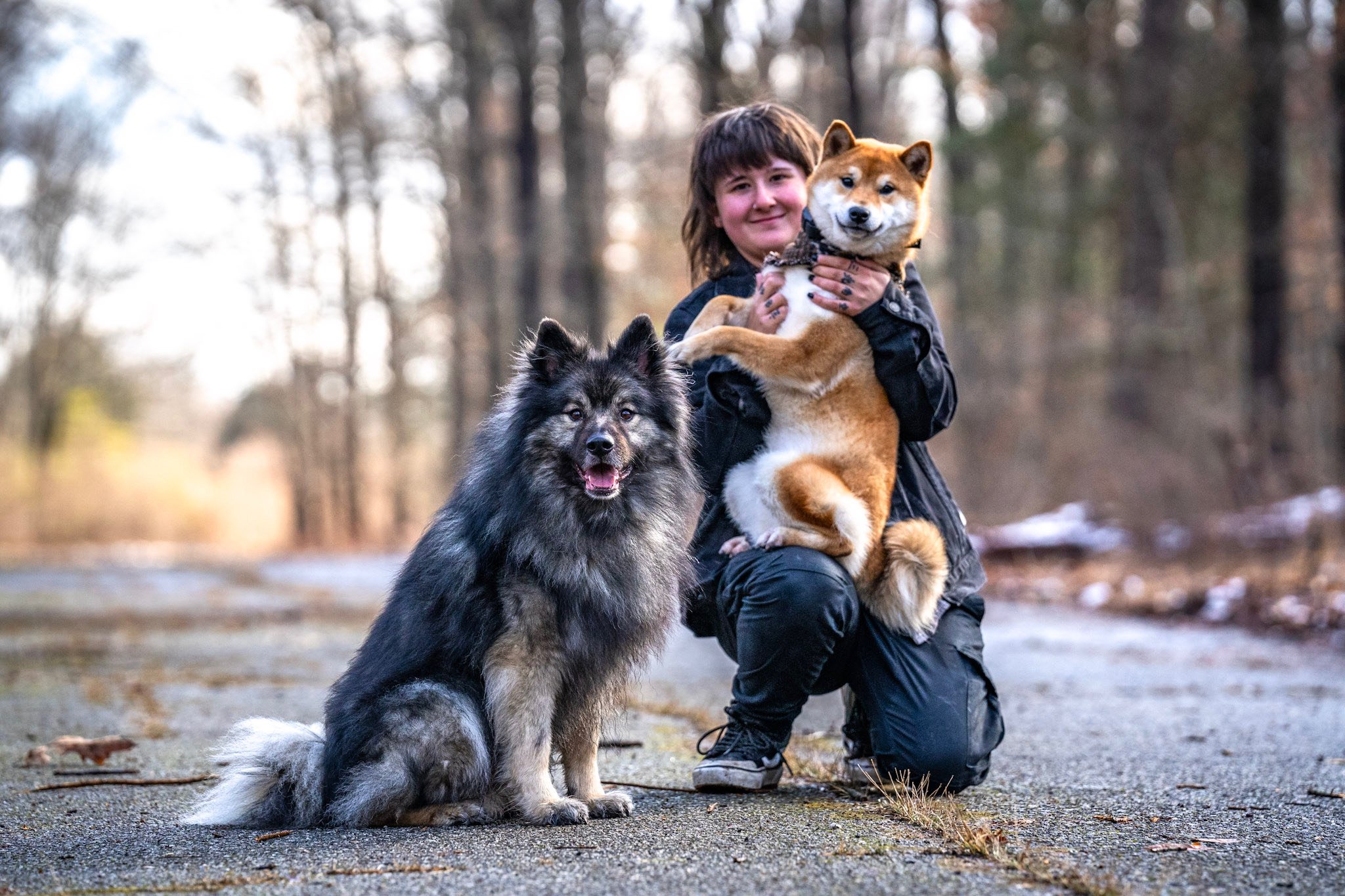 A woman kneeling on a forest path holding a Shiba Inu dog, with a Corgi sitting in front of her and looking at the camera. The background features bare trees and a slightly snowy ground.