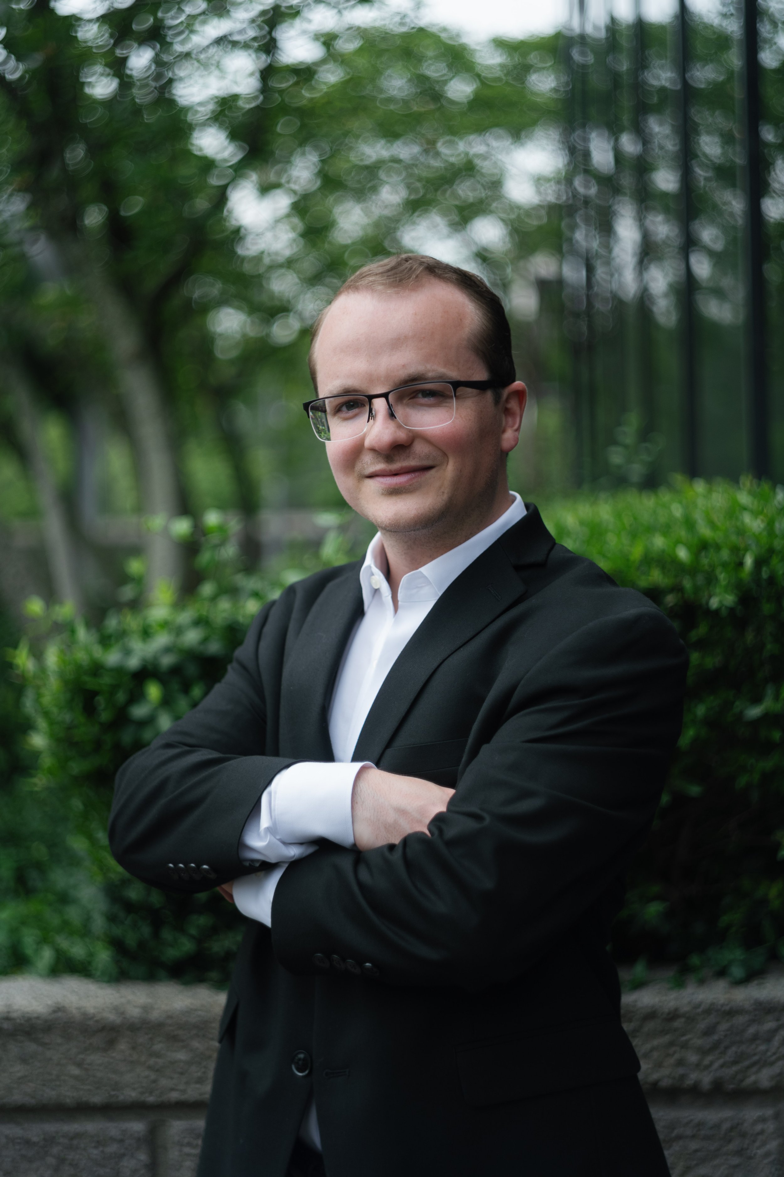 A man in a black suit with white shirt and glasses standing outdoors with arms crossed, background of greenery and blurred trees.