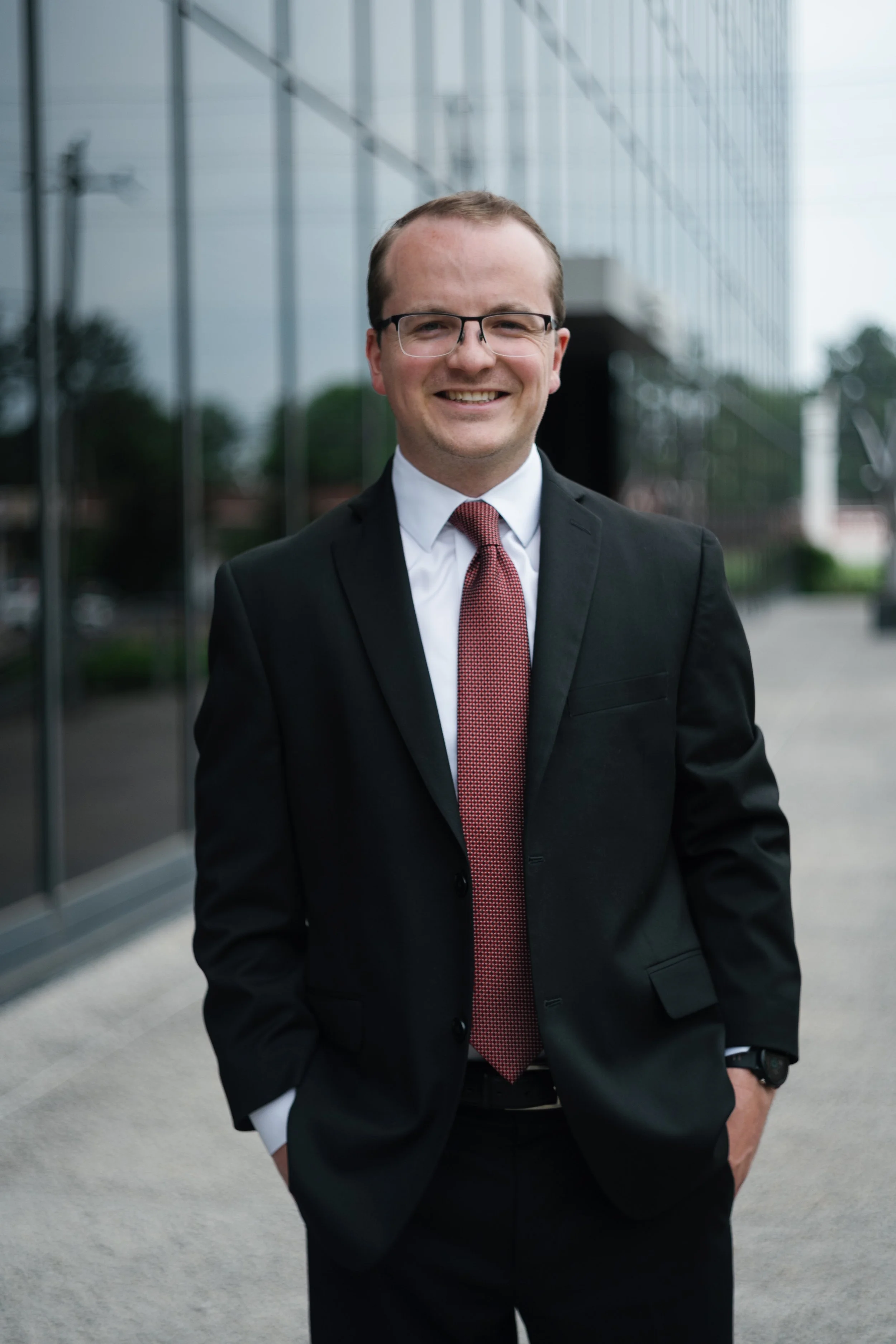 A man in a black suit and red tie standing outdoors in front of a reflective glass building, smiling with his hands in his pockets.