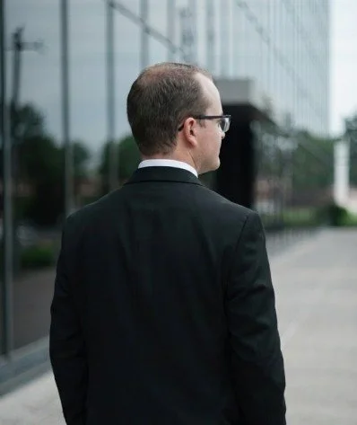 A man in a business suit wearing glasses stands outdoors near a modern glass building, facing away from the camera.