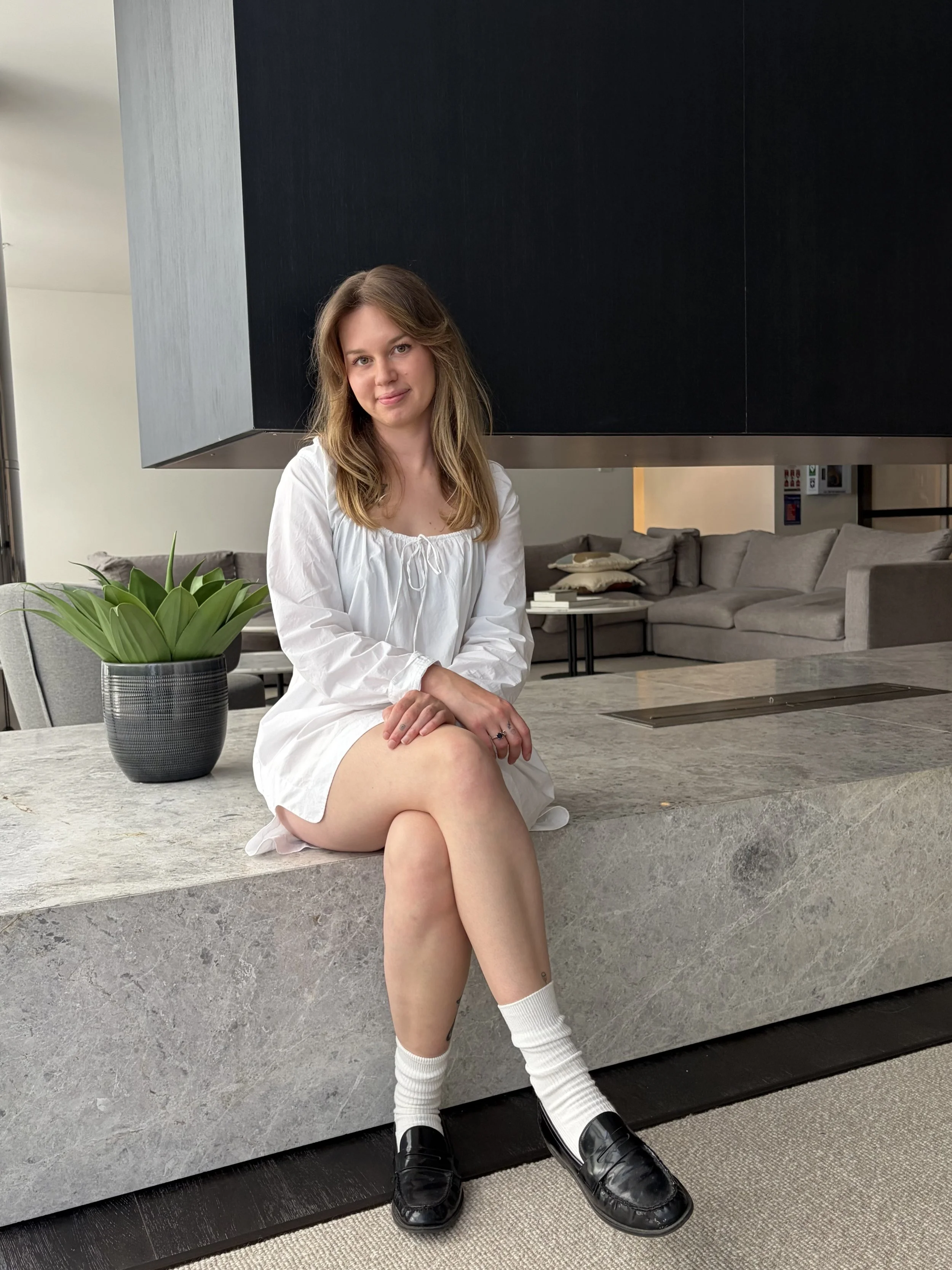 A young woman sitting on a marble ledge in a modern living space, wearing a white dress, black loafers, and white socks, with a potted plant beside her, and a gray sofa with pillows in the background.