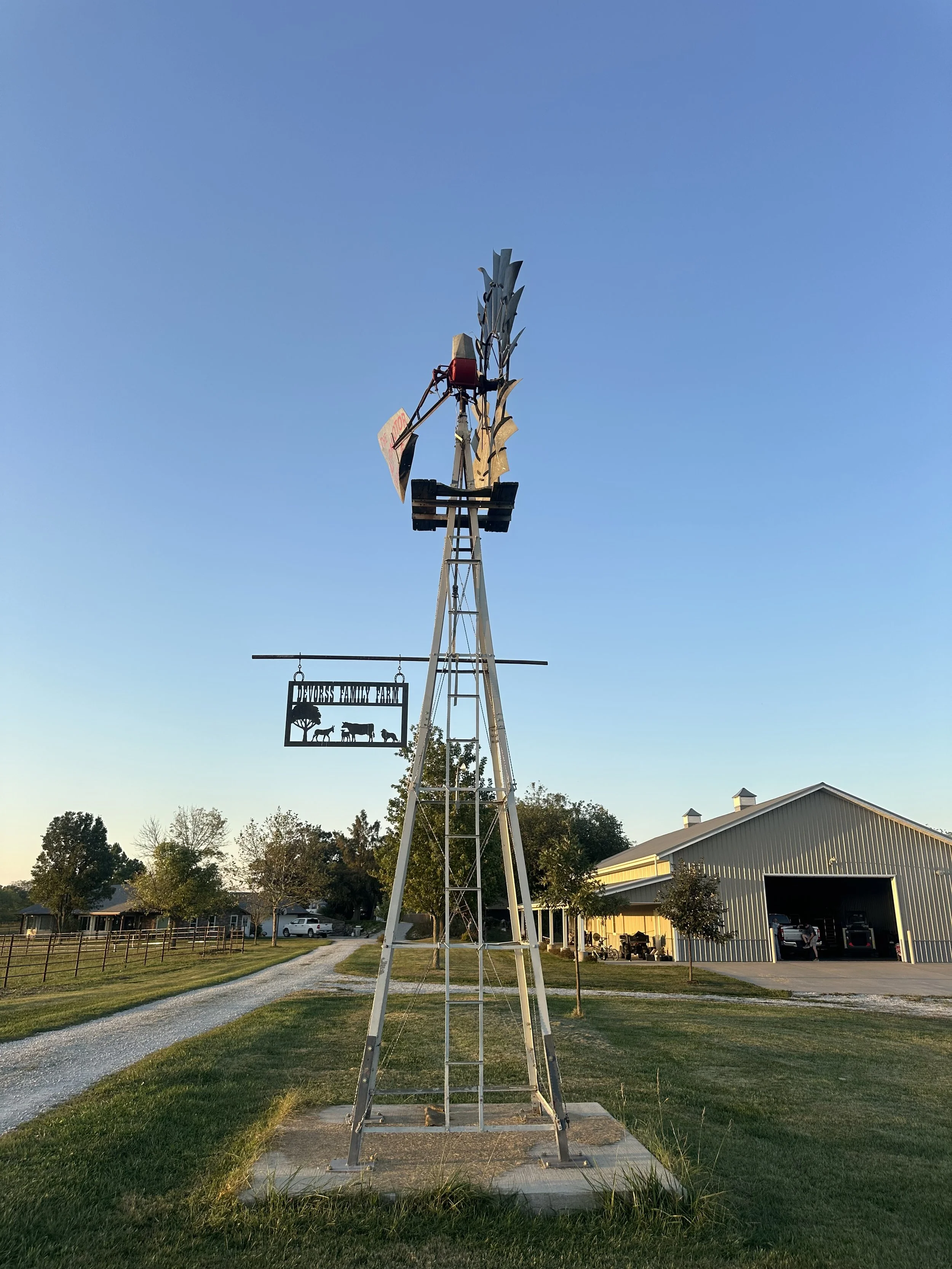 A tall metal windmill with a blade at the top, set against a clear blue sky. There is a sign hanging from a horizontal pole reading 'Hawks Family Farm' with silhouettes of farm animals. In the background, there is a large metal barn with some cars and trees nearby.