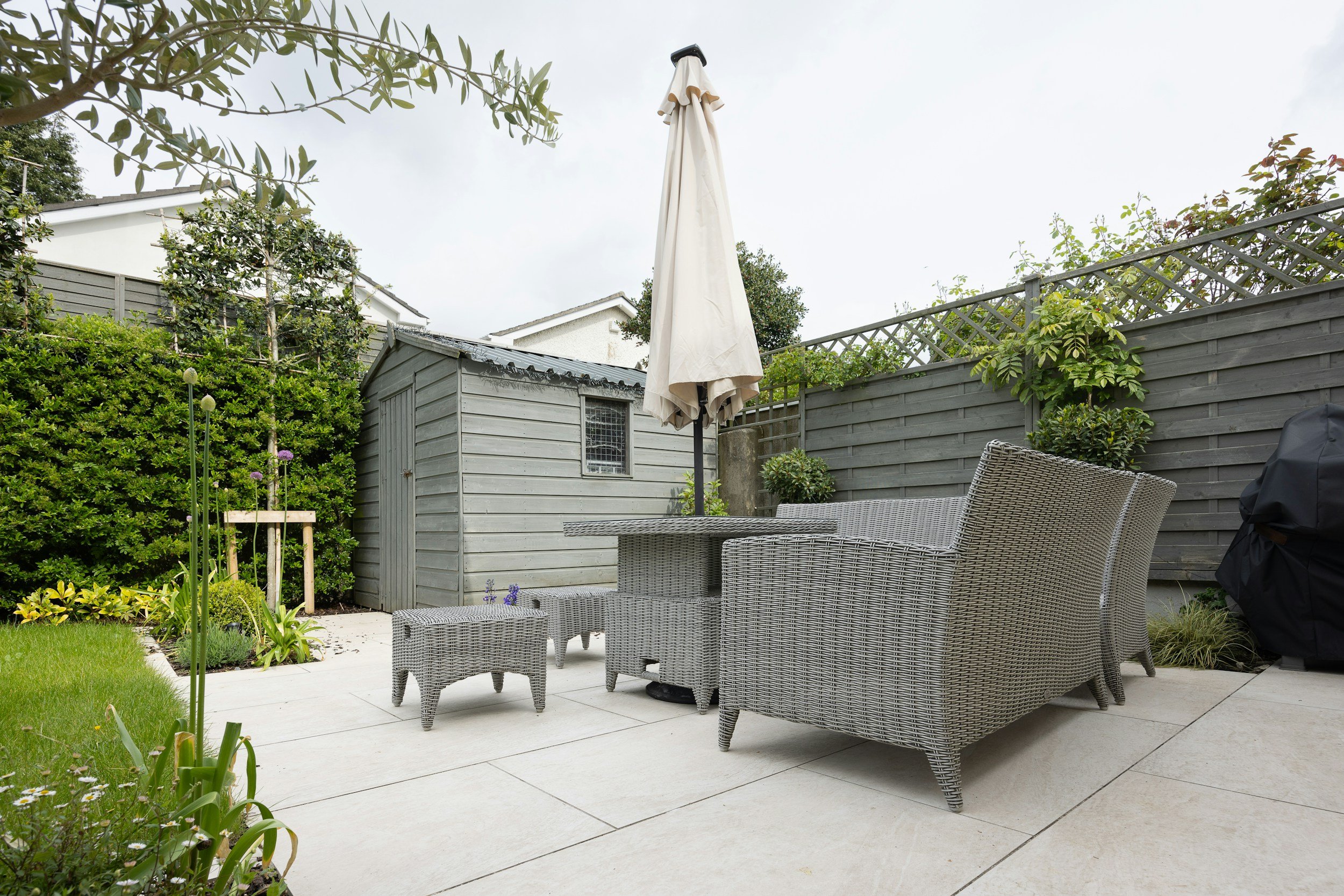 Patio area with patio furniture, including a wicker sofa, chairs, a small table, an umbrella, and a garden shed surrounded by plants and a wooden fence.