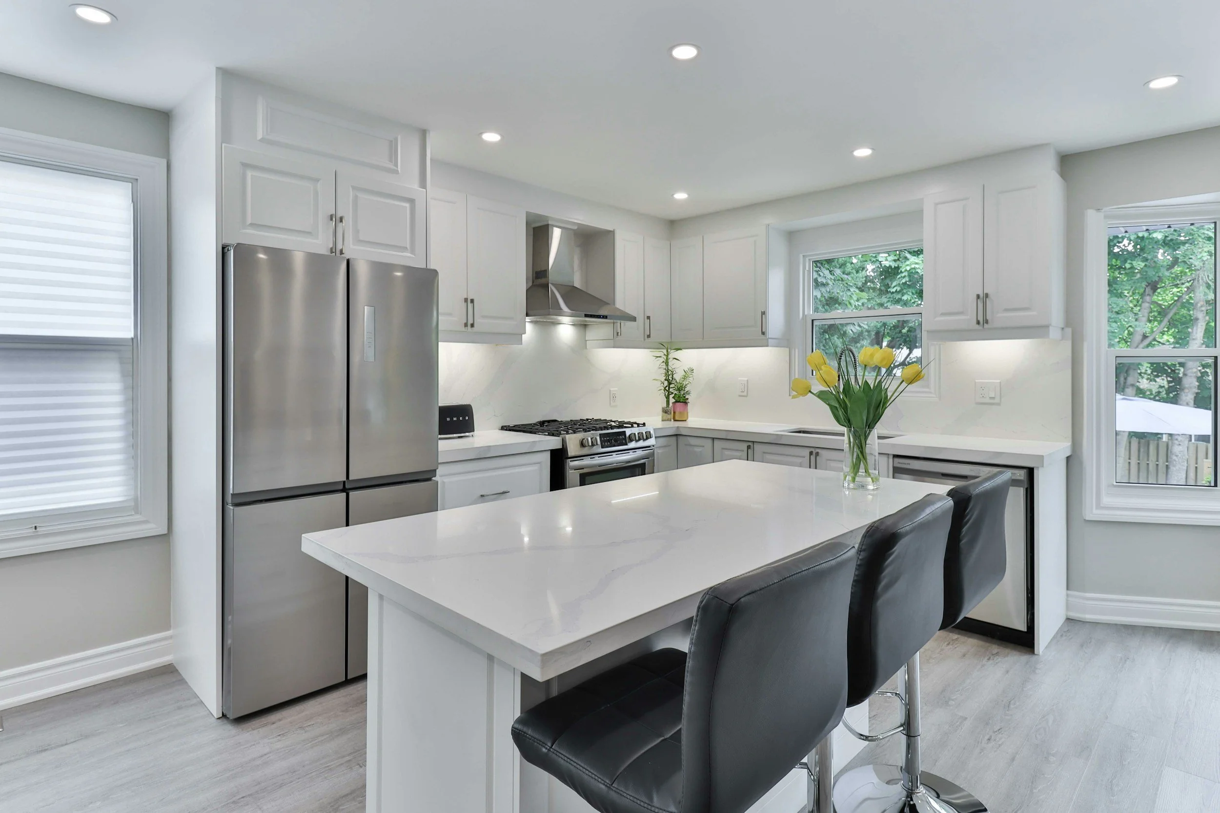 Modern kitchen with white cabinets, stainless steel refrigerator, gas stove, white countertops, and black bar stools with a vase of yellow tulips on the island.
