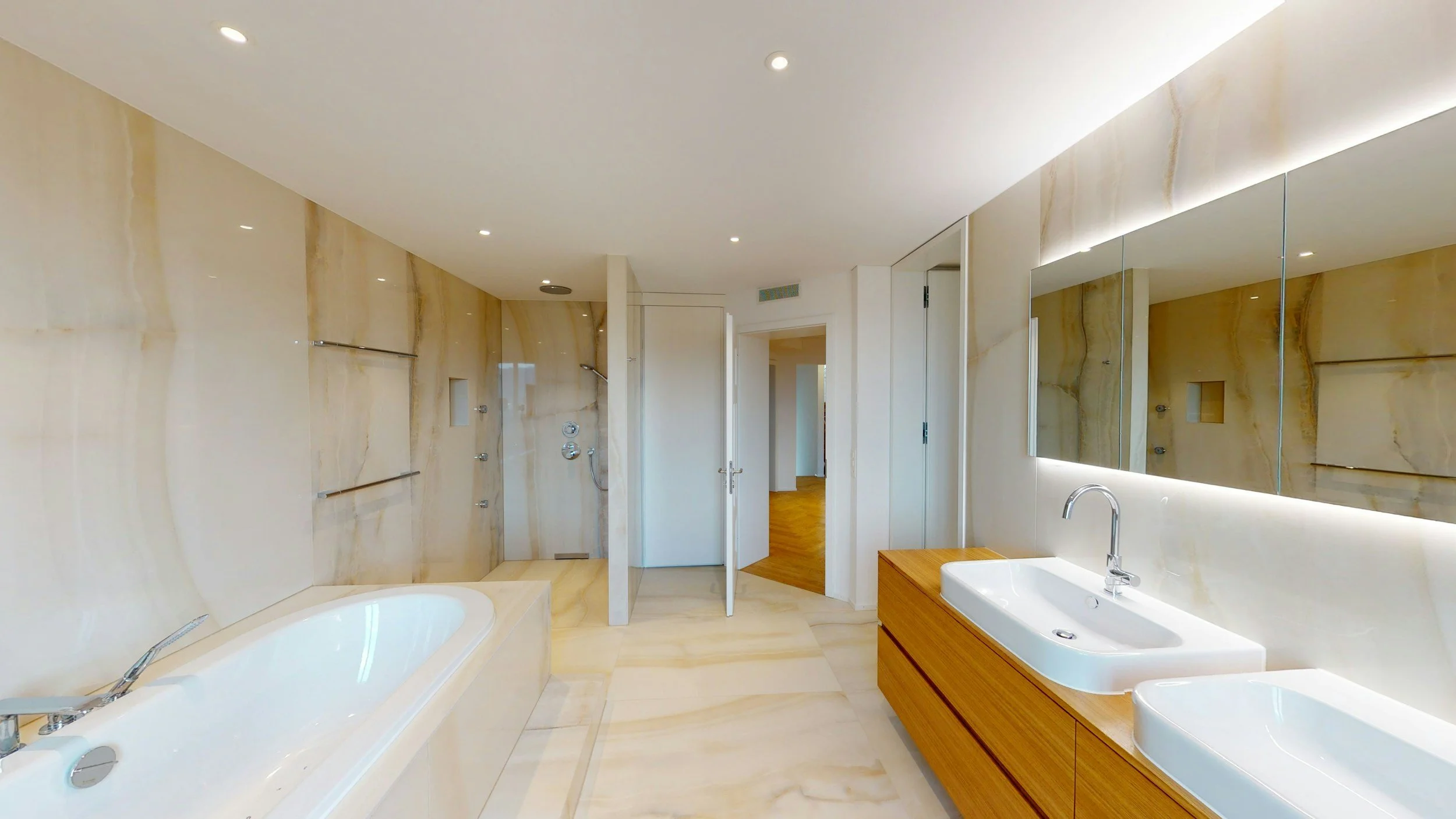 Modern bathroom with marble walls and floor, a built-in bathtub, a wooden vanity with double sinks, and a large mirror with backlighting.