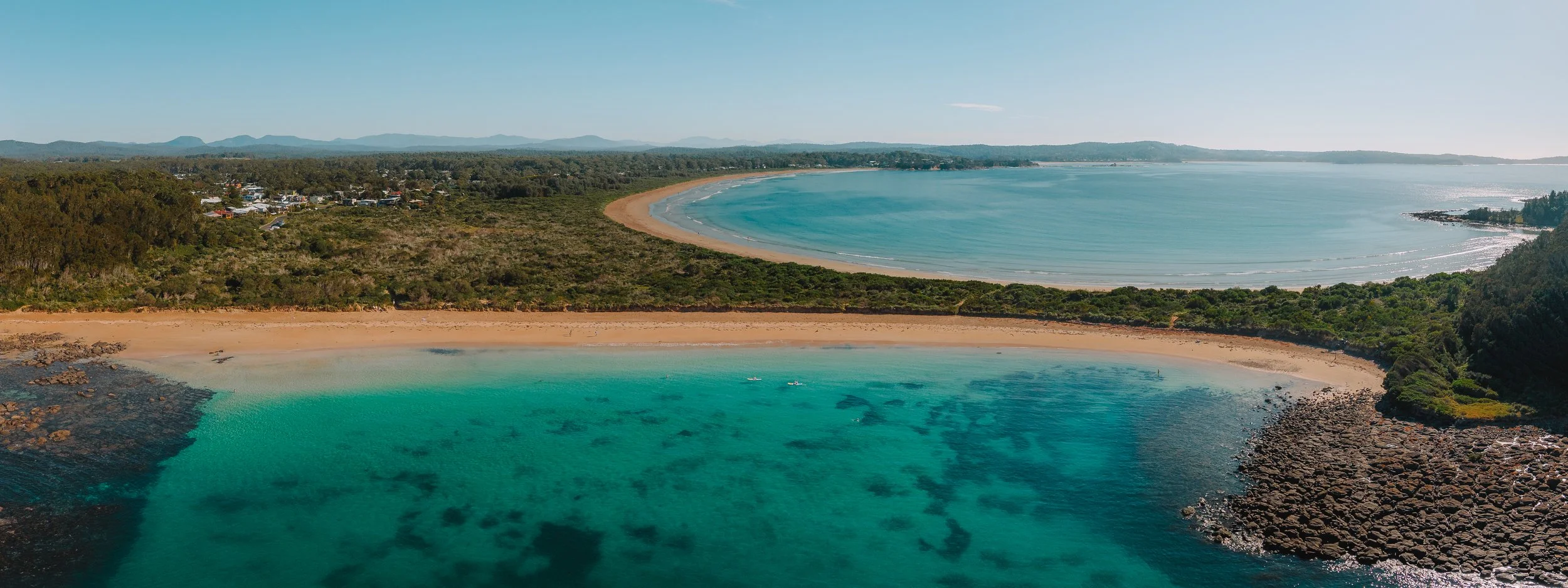 Aerial view of a coastal area with turquoise water, a sandy beach, green trees, and distant mountains under a clear blue sky.