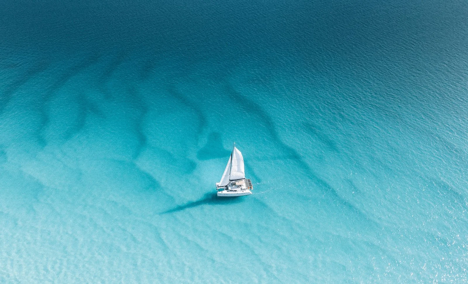 An aerial view of a sailboat with white sails on clear blue water, casting a shadow on the water.