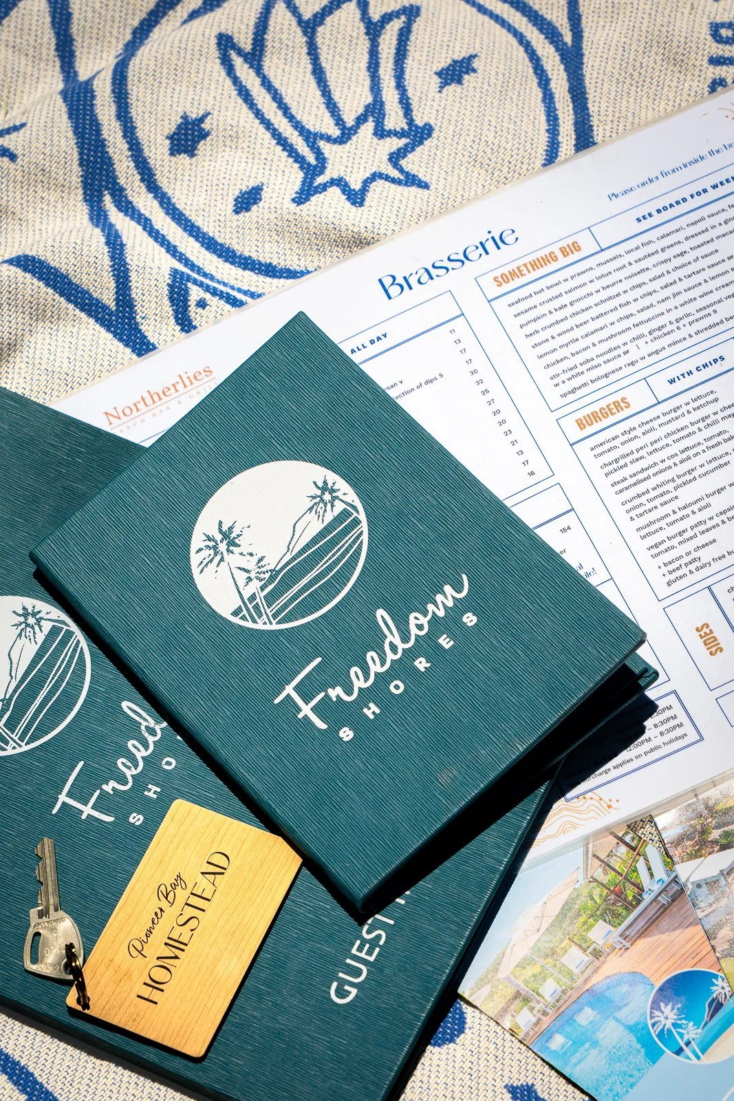 Brochure for Freedmen Shores with palm trees and mountains logo, placed on a blue folder, alongside a hotel keycard labeled 'Point Bay Homestead,' and a menu from a restaurant named Brasserie, all on a table with a blue and white cloth.