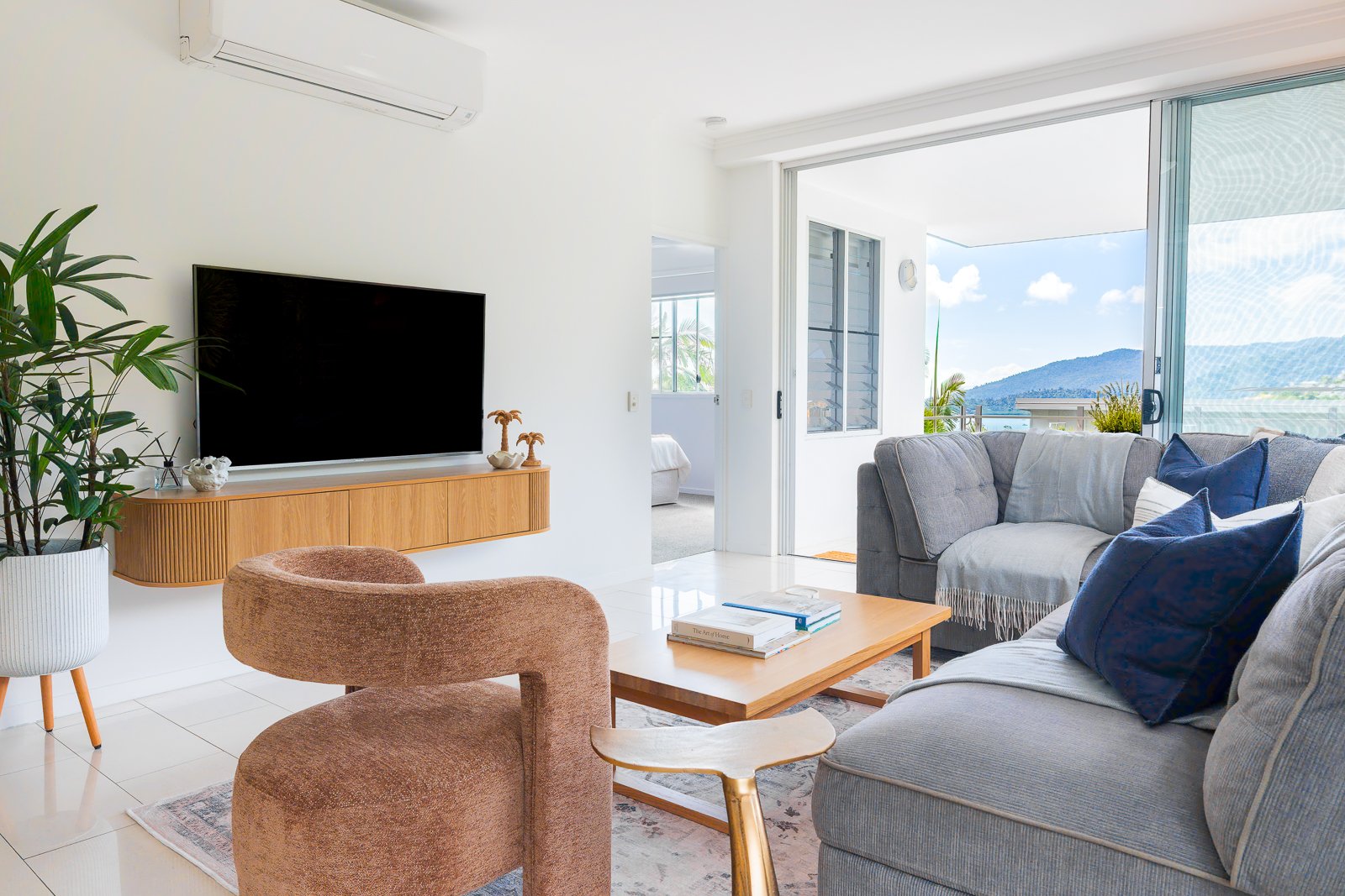 Living room with a large TV, pink armchair, gray sofa with navy and white pillows, wooden coffee table with books, indoor plants, sliding glass door leading to balcony with view of mountains and blue sky.