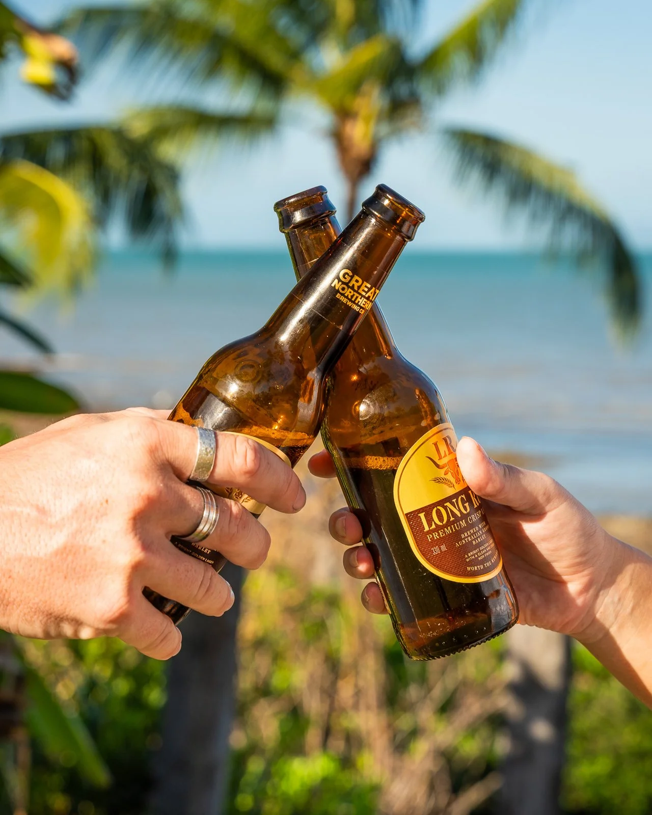Two people clinking brown glass beer bottles labeled Great Northern Brewing Co. and Long White Beer in front of a tropical beach background with palm trees and ocean