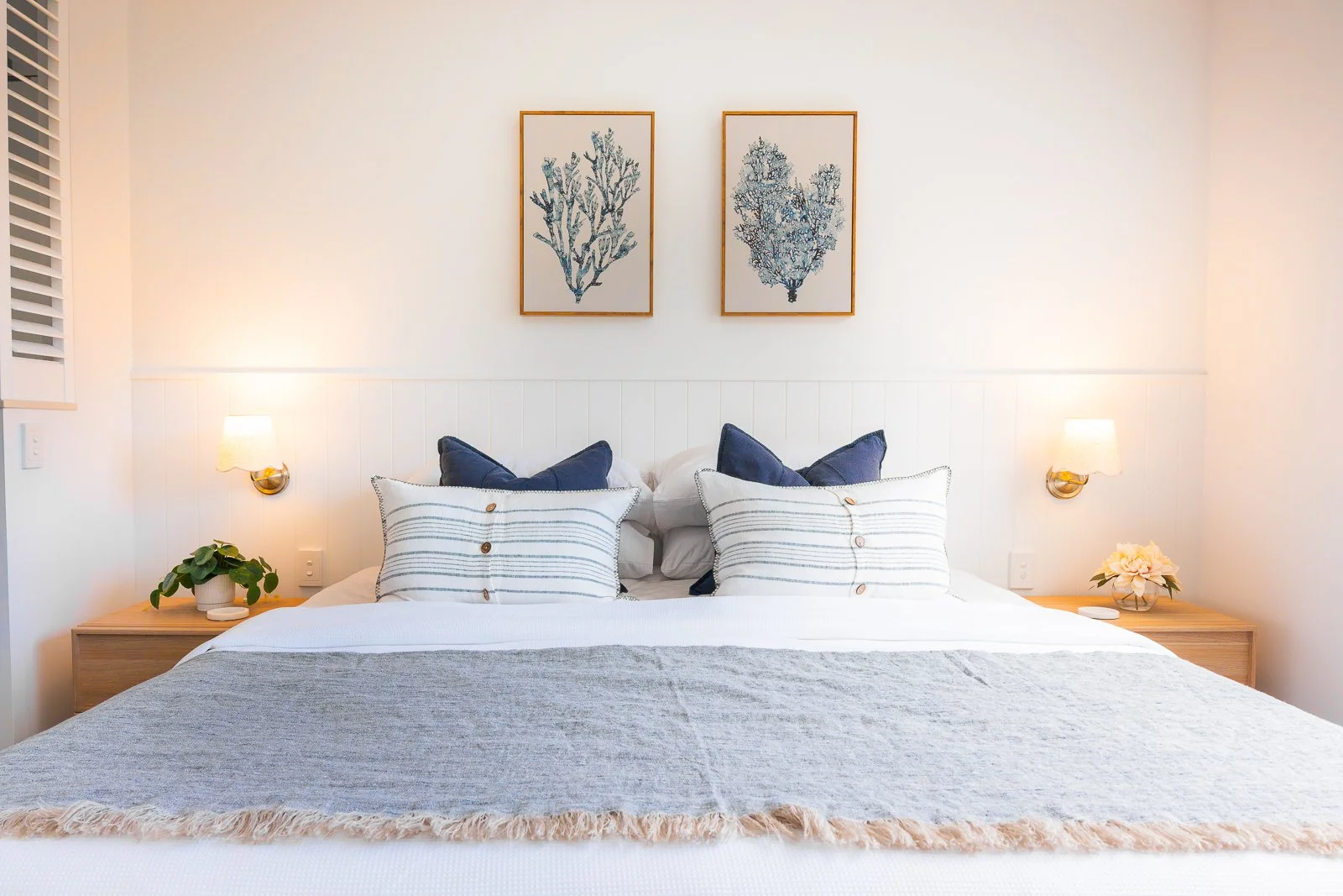 A neatly made bed with white and navy pillows, a light gray runner, and two wooden nightstands with lamps, a plant on the left, and flowers on the right, against a white wall with two framed botanical prints.