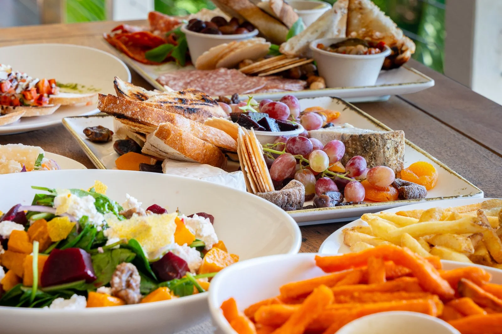Assorted dishes on a table, including a cheese and fruit platter, a salad with beets and greens, a plate of fries, and a pasta dish, with an additional platter of bread, grapes, olives, and meats in the background.