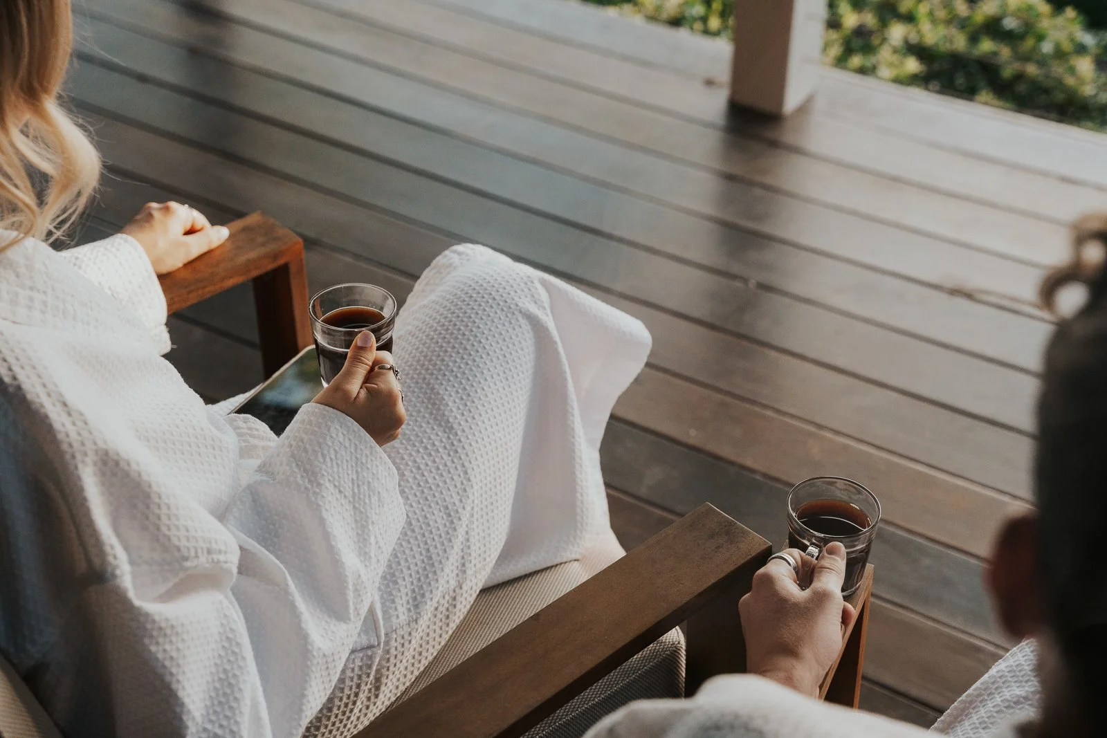 Two women in white robes relaxing on a porch, each holding a glass cup of coffee or tea.