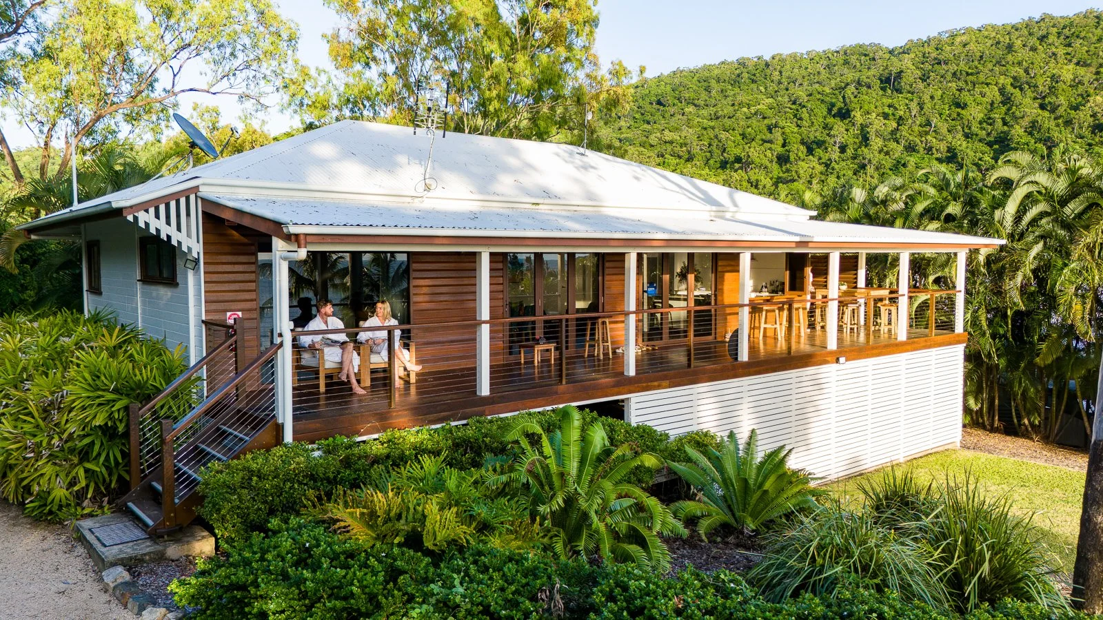 A modern elevated house with a white metal roof and extensive wooden deck, surrounded by lush tropical greenery and trees in a hilly landscape. Two people are sitting and talking on the deck, which has outdoor seating and barstools.