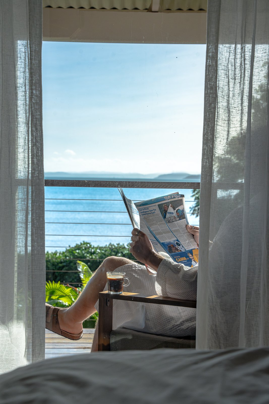 Person sitting on a balcony with a view of the ocean, reading a magazine, with a cup of coffee on a small table.