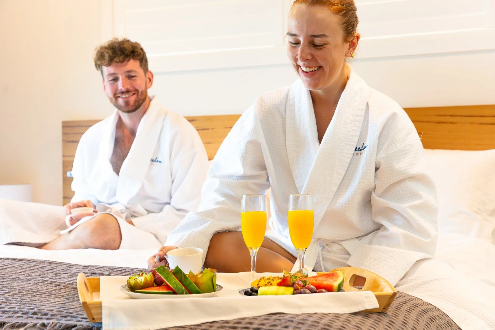 A smiling woman and man in bathrobes sitting on a bed with breakfast tray featuring fruit, orange juice, and coffee in a hotel room.