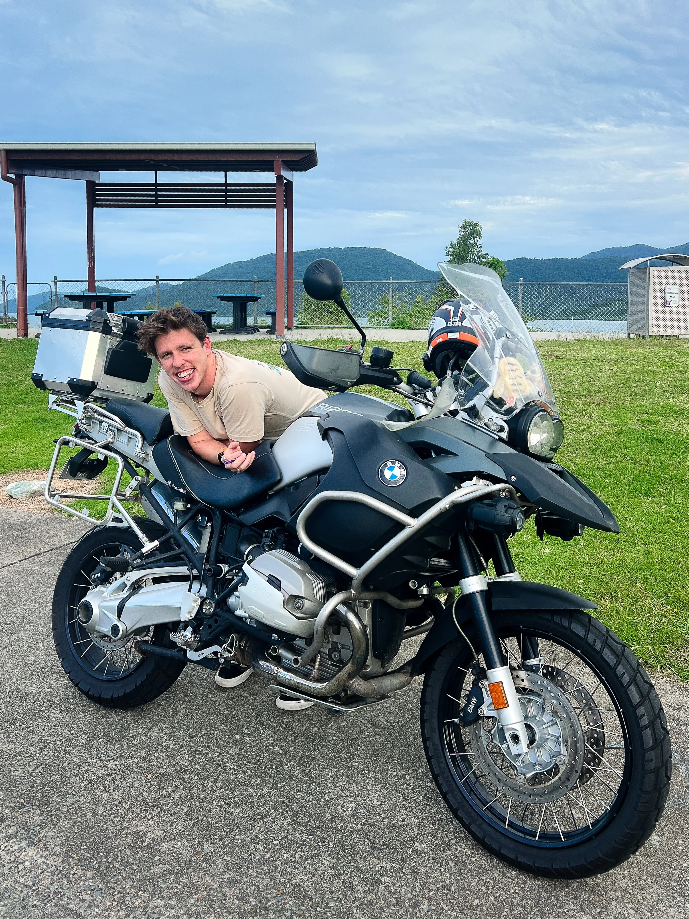 A smiling man leaning on a black and silver BMW adventure motorcycle with luggage, parked on a paved surface near a grassy area with picnic tables and a mountain view in the background.