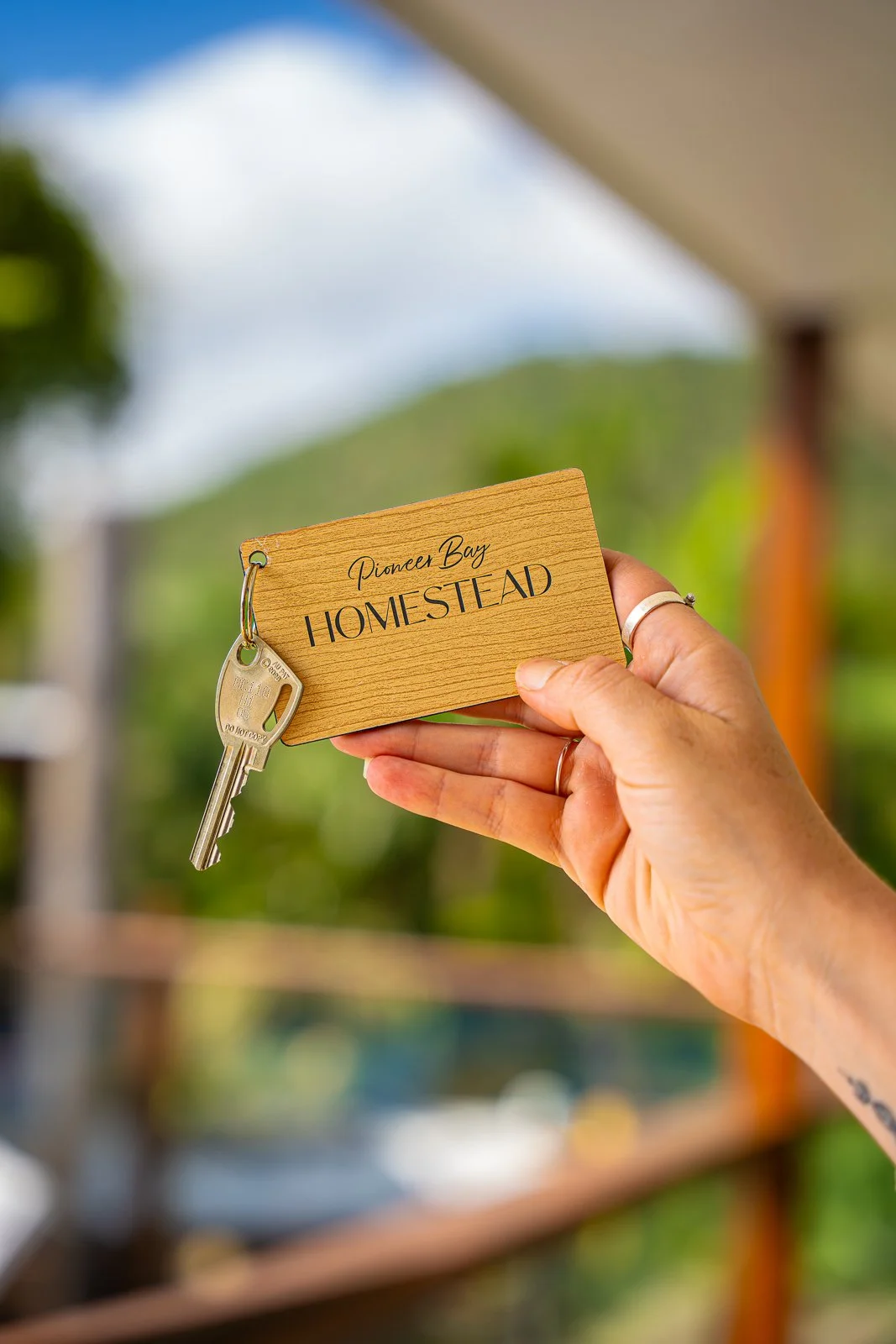 A hand holds a wooden keychain with the words 'Pioneer Bay Homestead' engraved on it, along with a silver key attached to the keychain. The background shows an outdoor setting with blurred greenery and a cloudy sky.