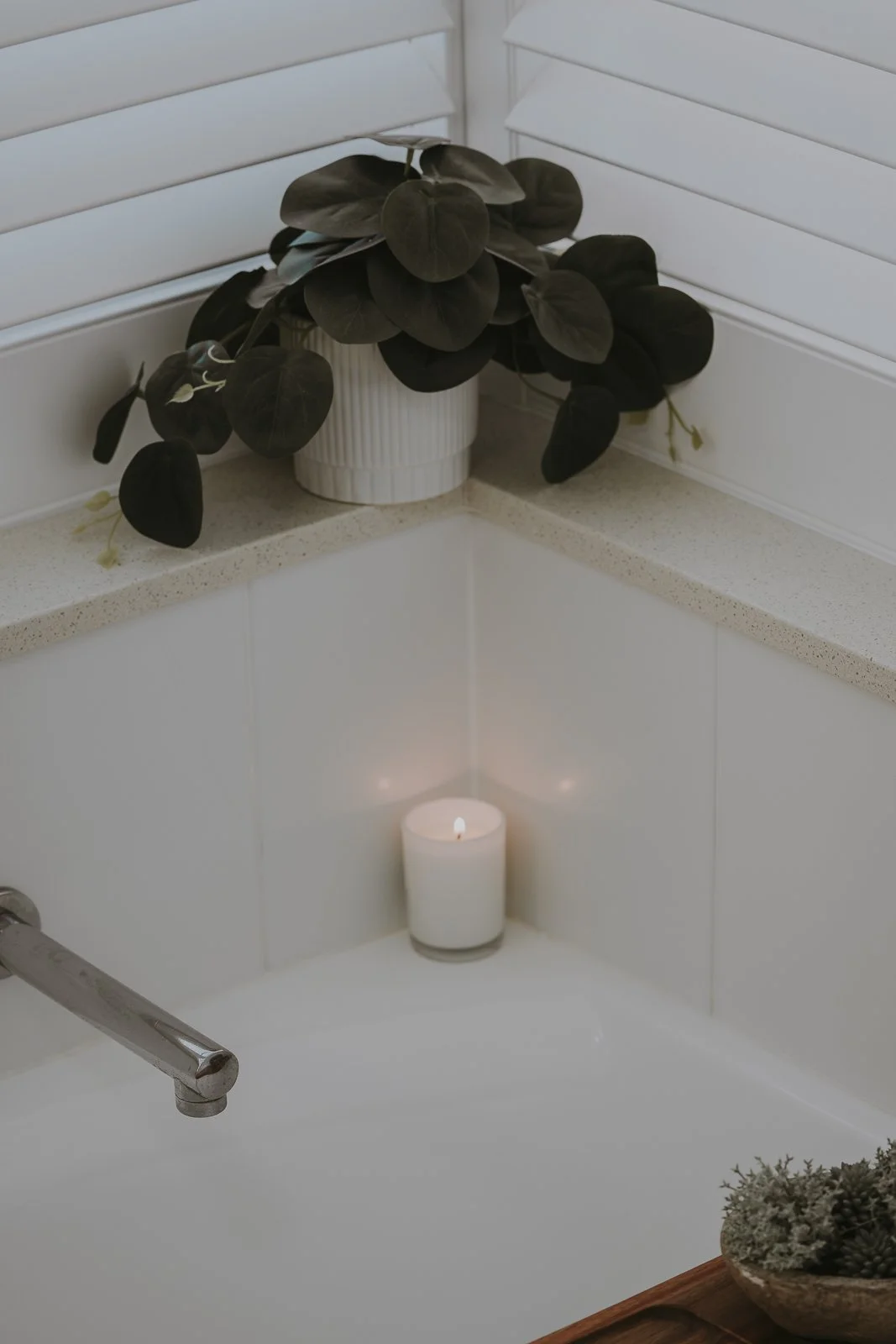 A white candle lit in a bathroom corner with a potted plant on a shelf above it.