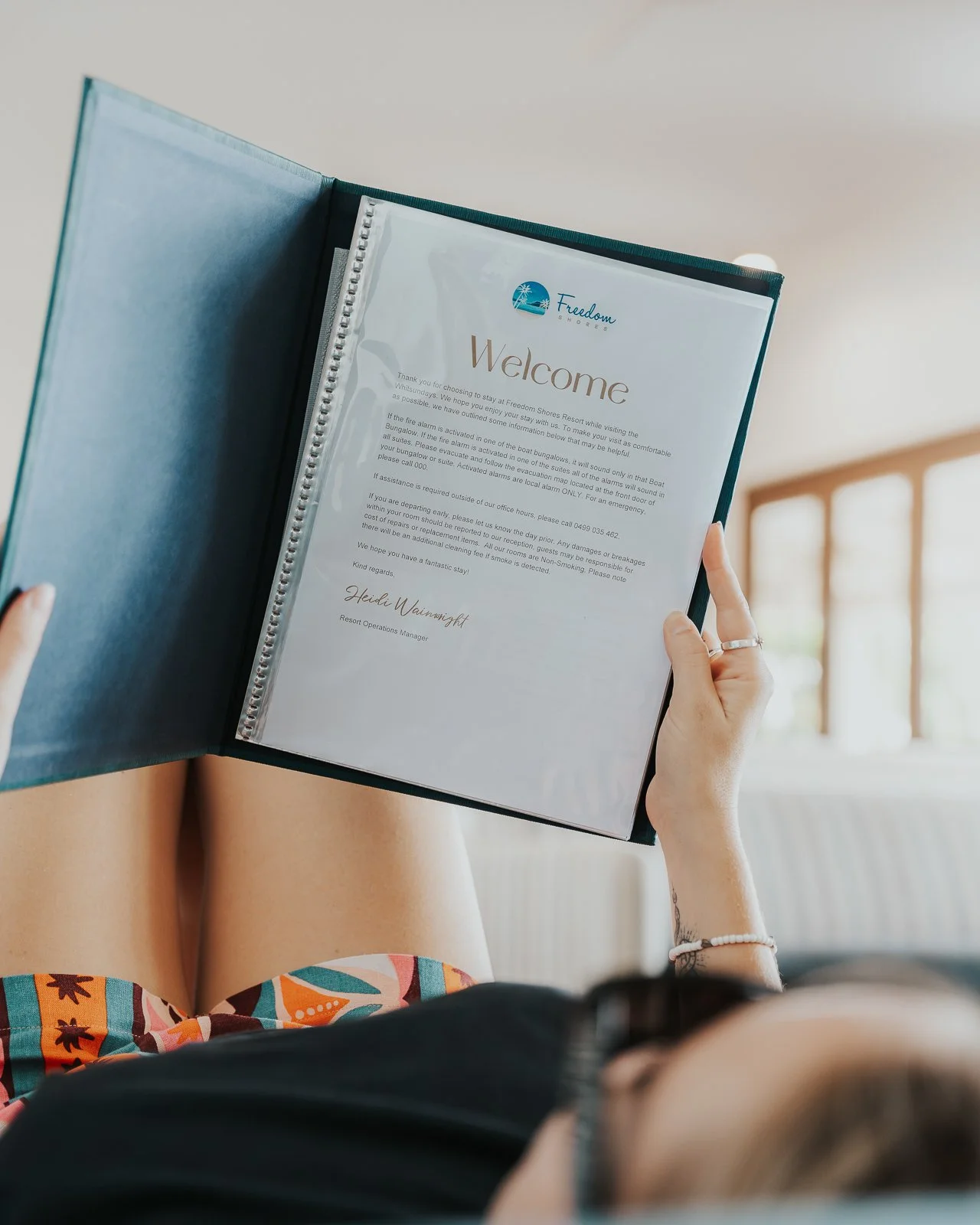 Person holding a welcome letter from Freedom Shores Resort, sitting with legs crossed, wearing a black top and colorful shorts, in a bright room with large windows.