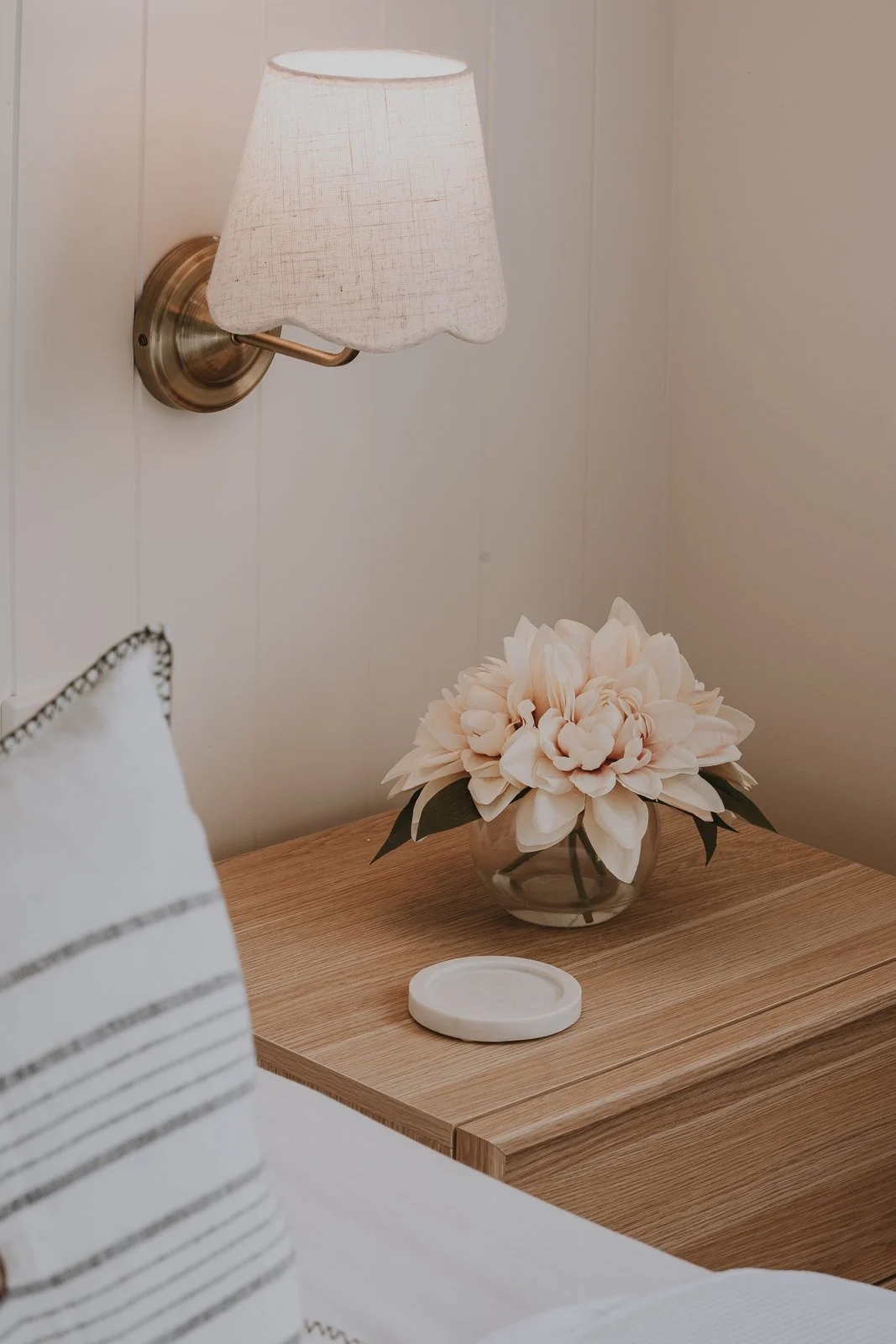 A bedroom bedside table with a floral vase, a white coaster, a wall-mounted lamp with a white fabric shade, and a pillow with black and white striped details.