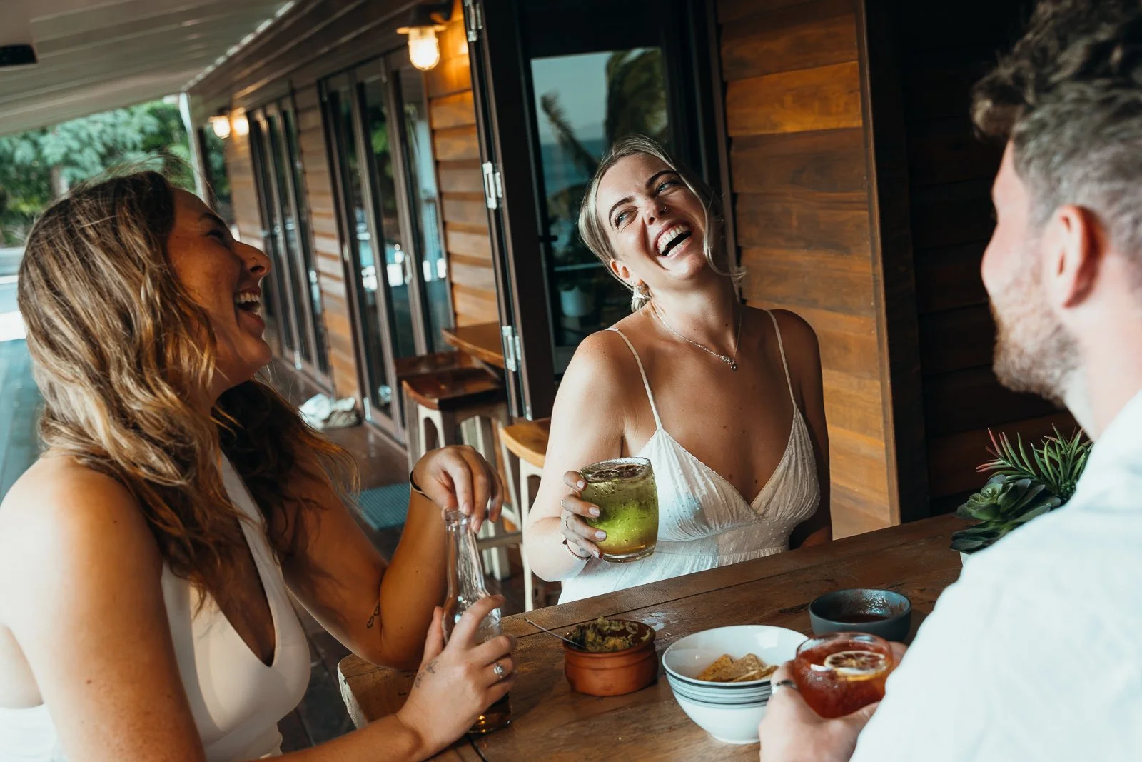 Three friends at a wooden bar or restaurant, smiling and laughing, with drinks and snacks on the table.