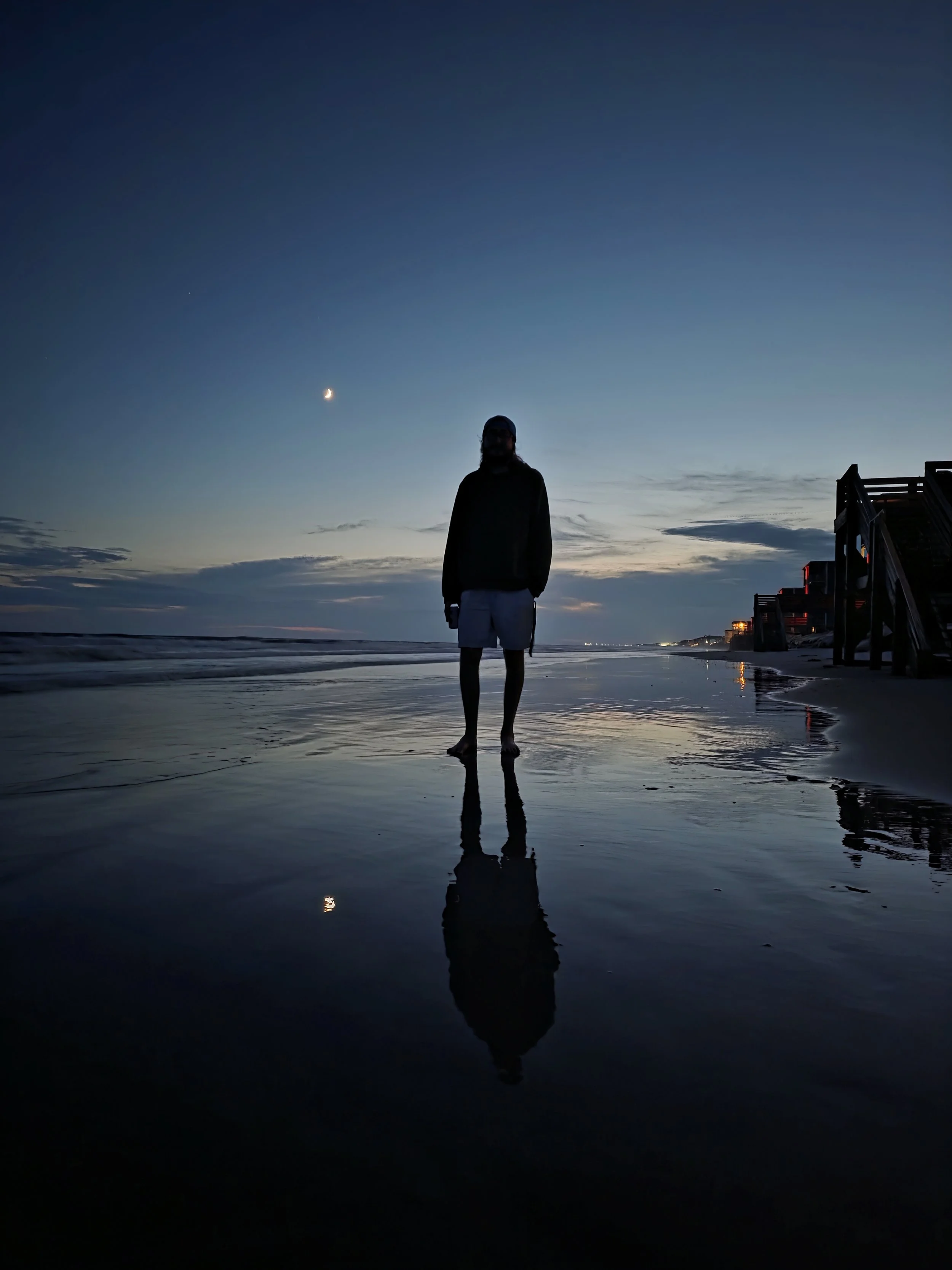 A person standing on a beach during sunset or dusk, with the moon and a star visible in the sky, and their reflection seen in the wet sand.